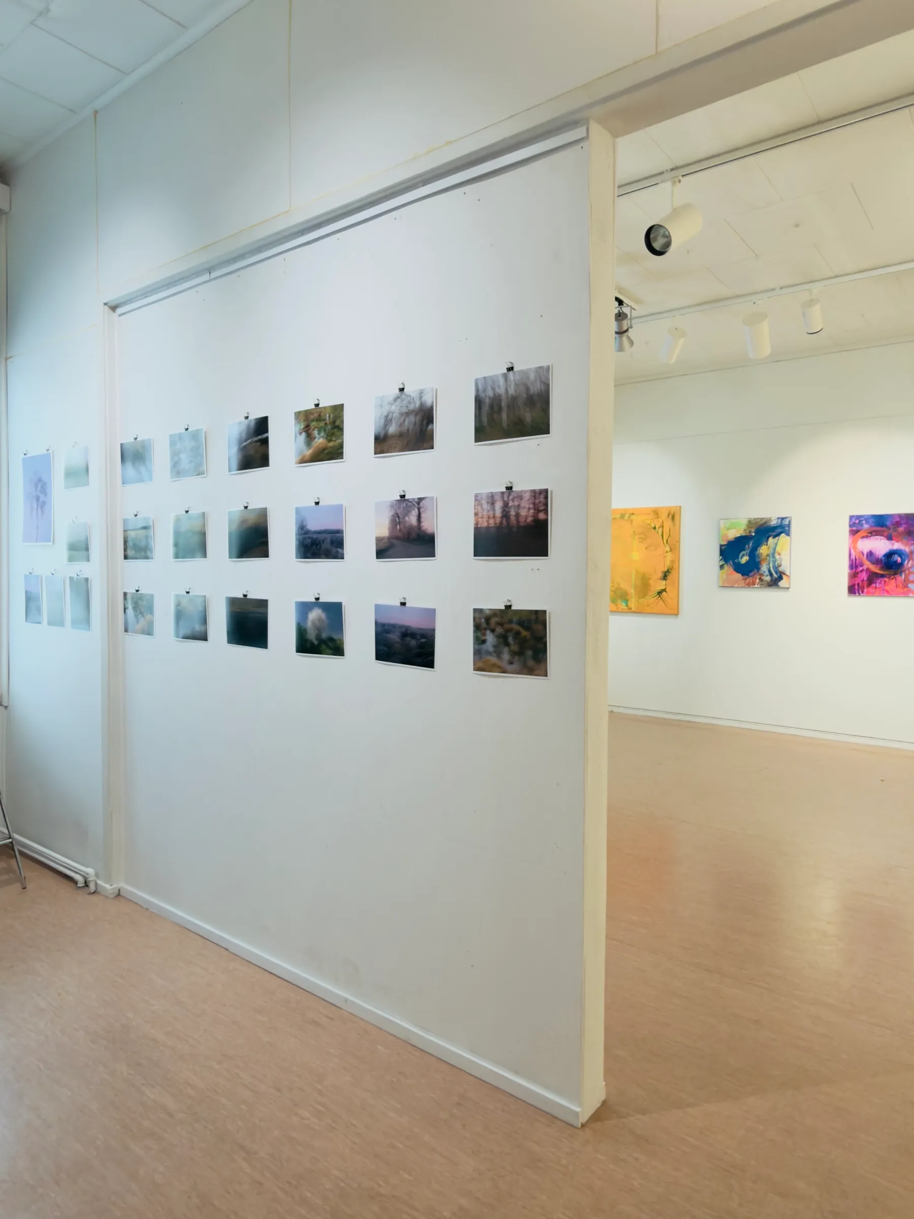 View through an exhibition room at Kaputa ry, photographic prints in the foreground, abstract paintings in the background