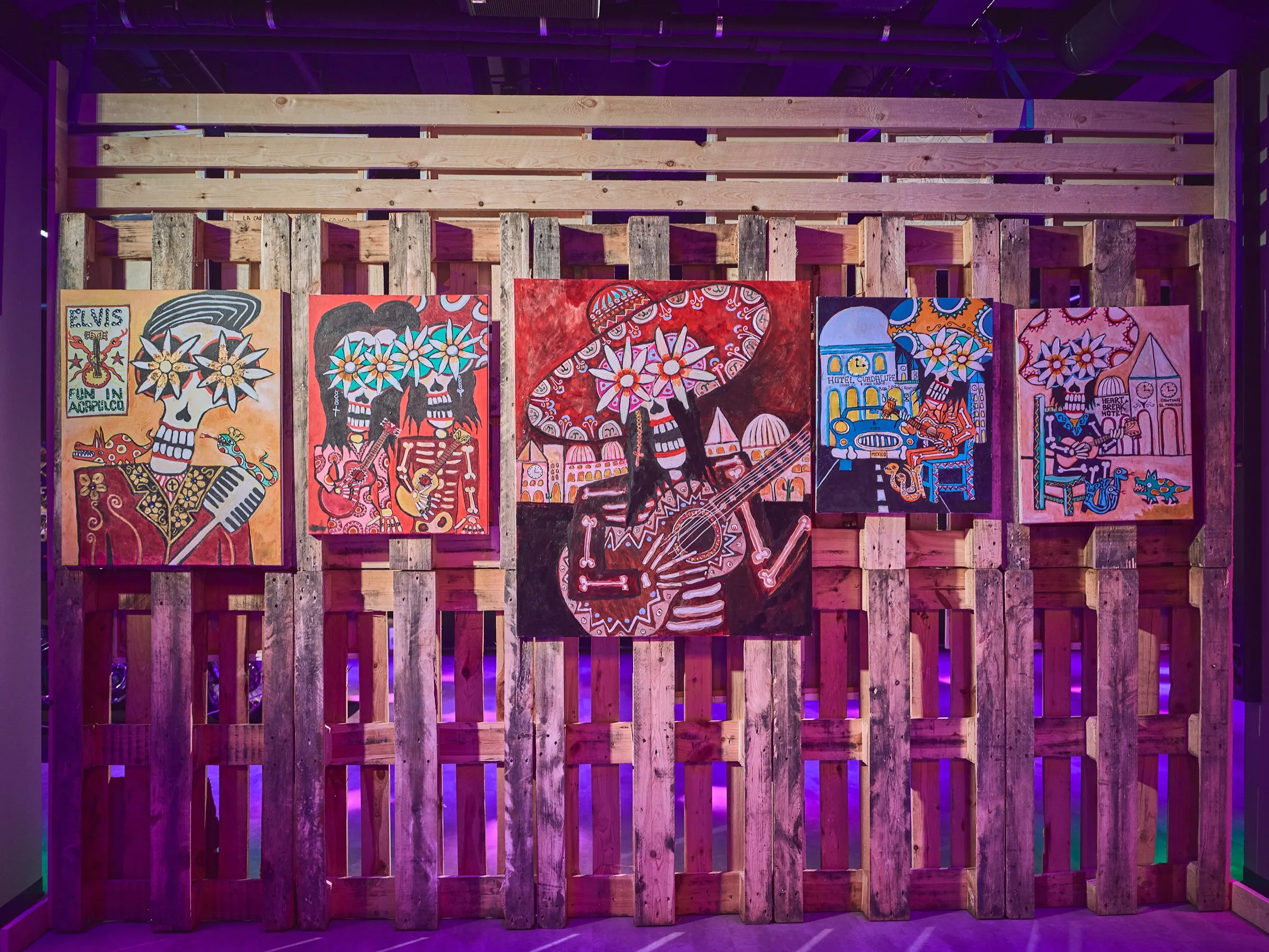 Wall of pallet wood hung with Day of the Dead paintings, a red picture with a dancing skeleton in the middle