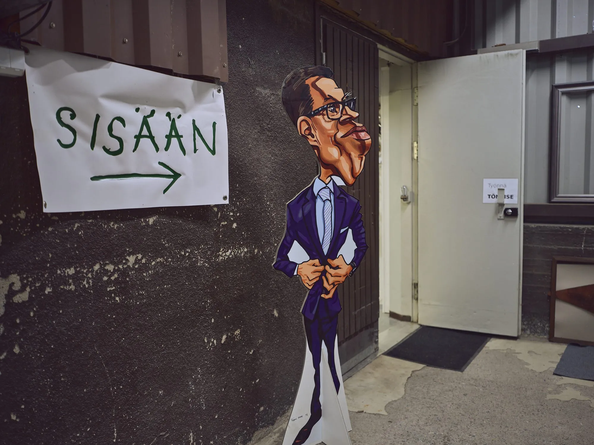 Three very different caricatures of Alexander Stubb side by side on a wall, as a playing card, as an oil painting, and as a comic couple