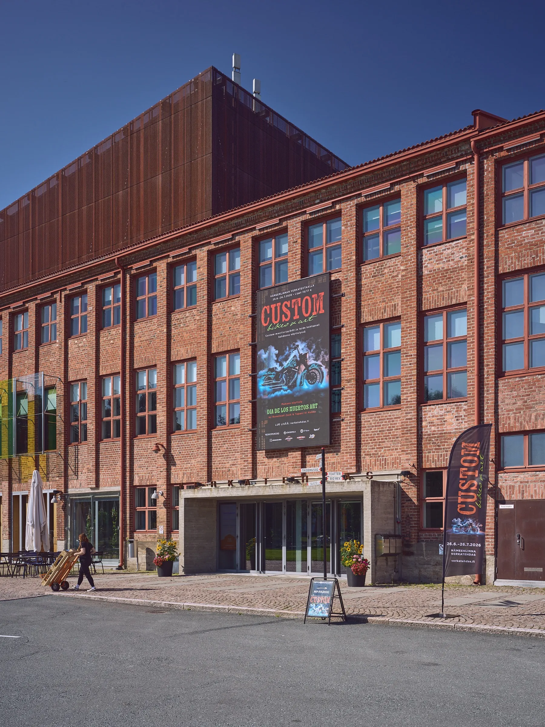 Brick façade in sunlight, a large poster for the Custom Bikes & Art exhibition on the wall, a banner by the entrance