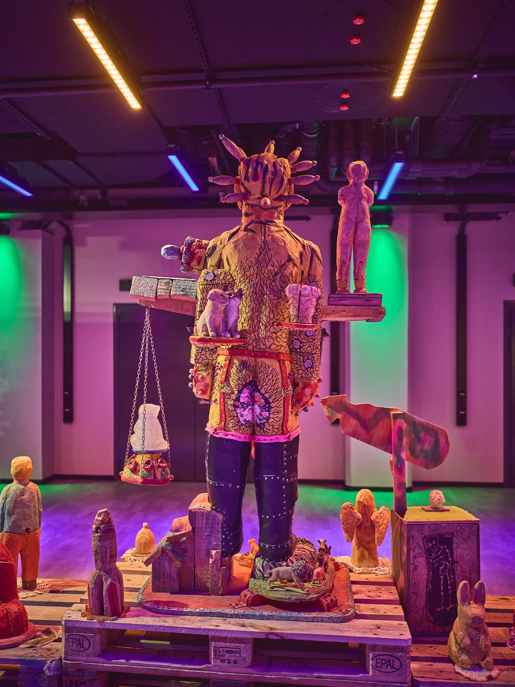 Tall figure with a feather headdress in front of a light wall, green and purple light