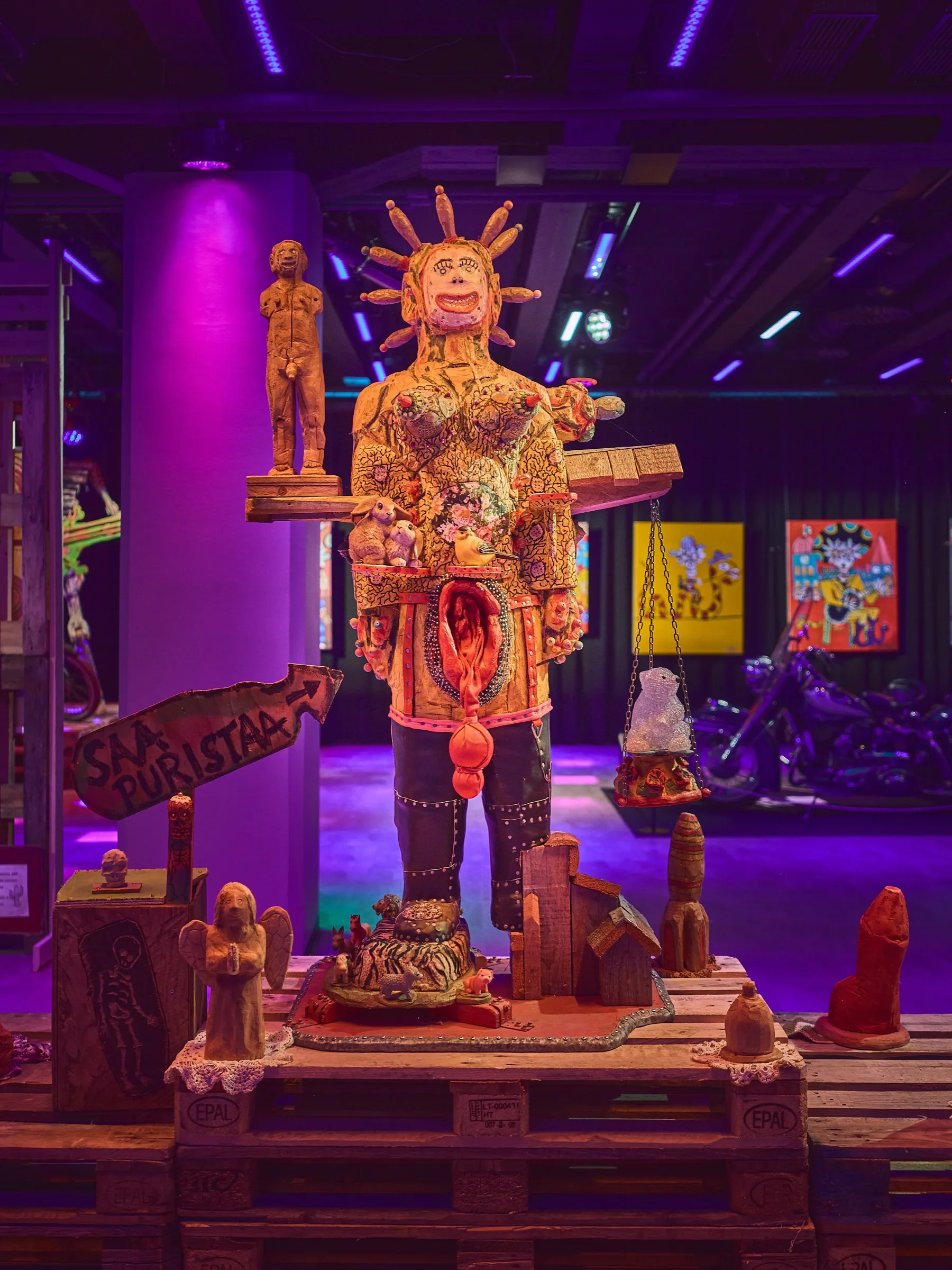 Man-high carved figure with a radiating headdress on a plinth, a motorcycle in the background