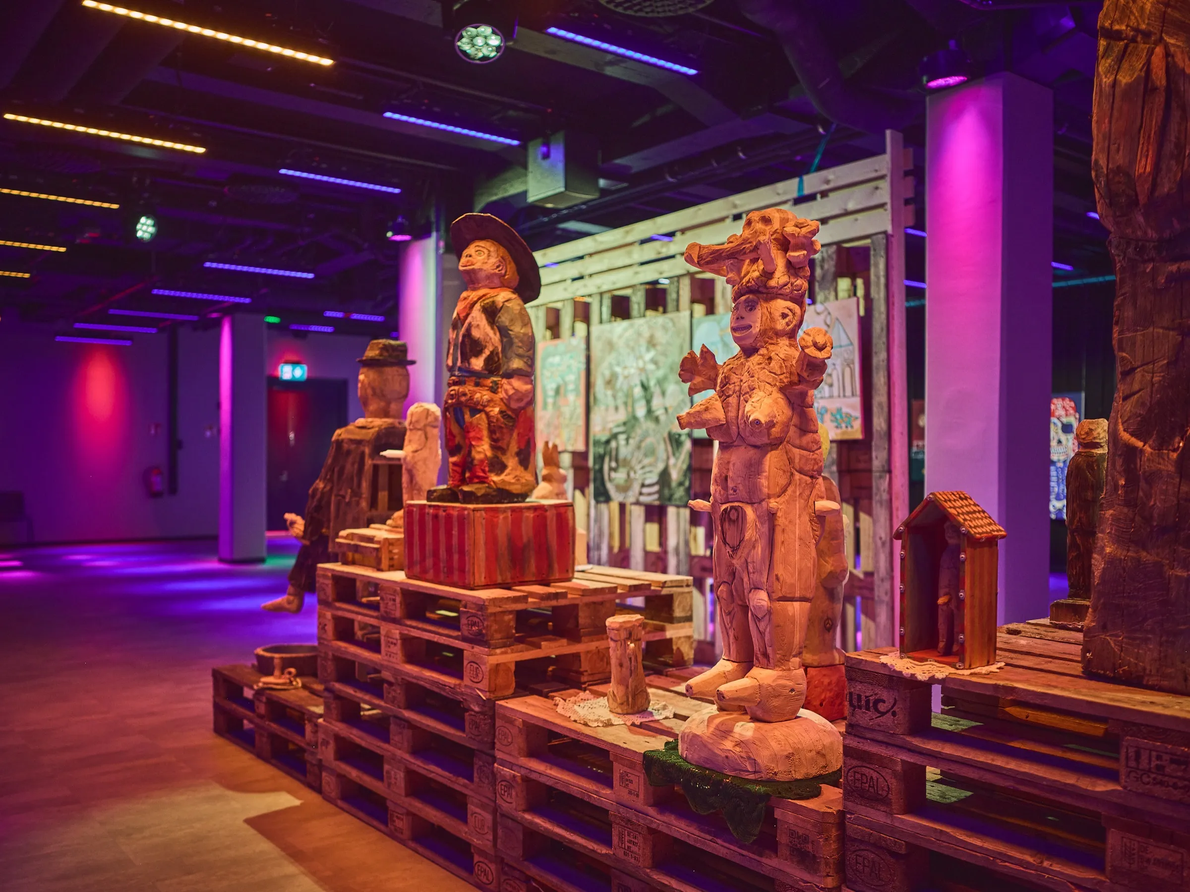 Group of tall wooden figures on pallets between purple-lit columns