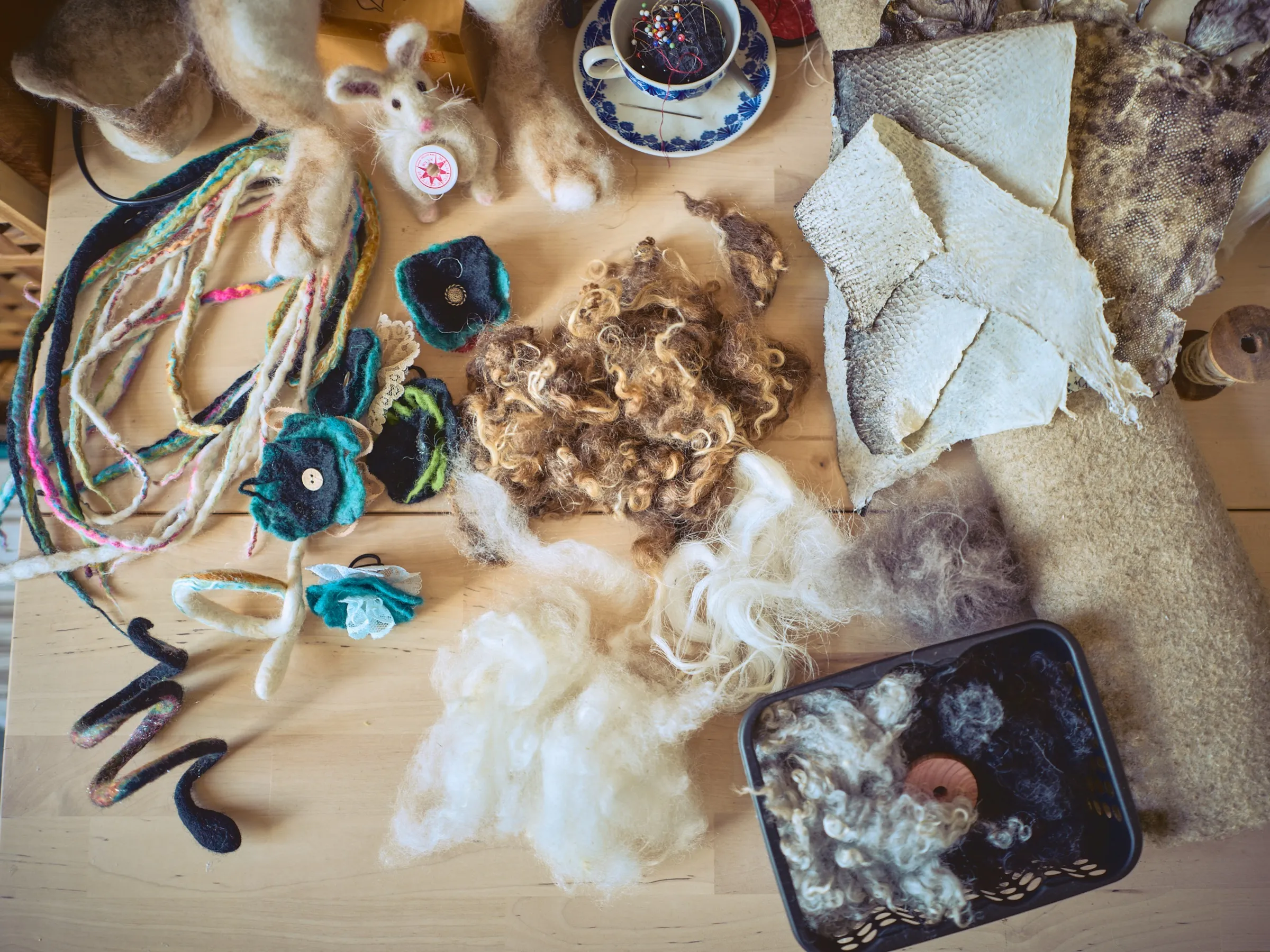 Work table from above: raw wool, felted flowers, cords, fish leather and a cup used as a pincushion