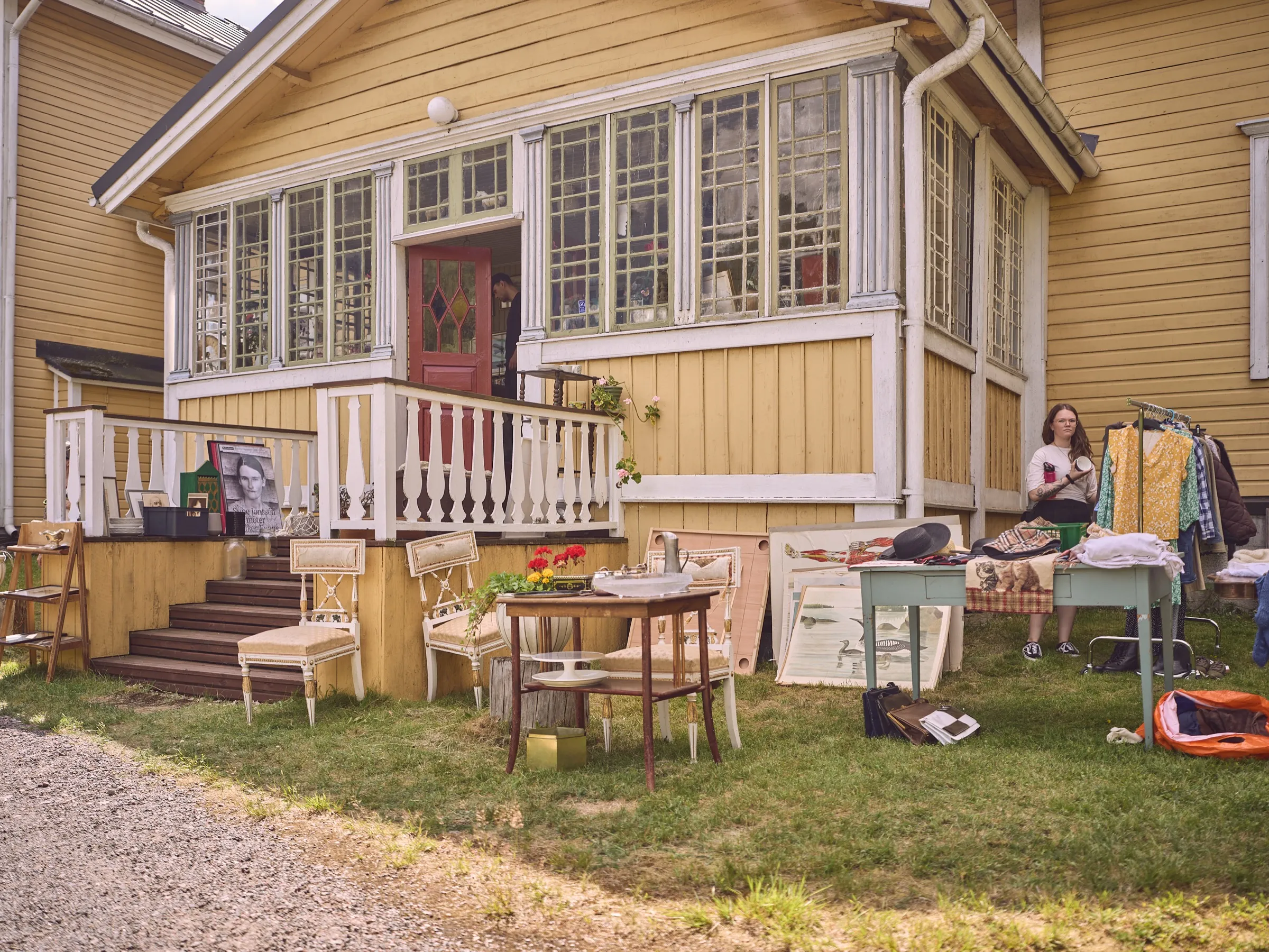 Yellow wooden house with veranda and steps, flea market furniture in the grass in front