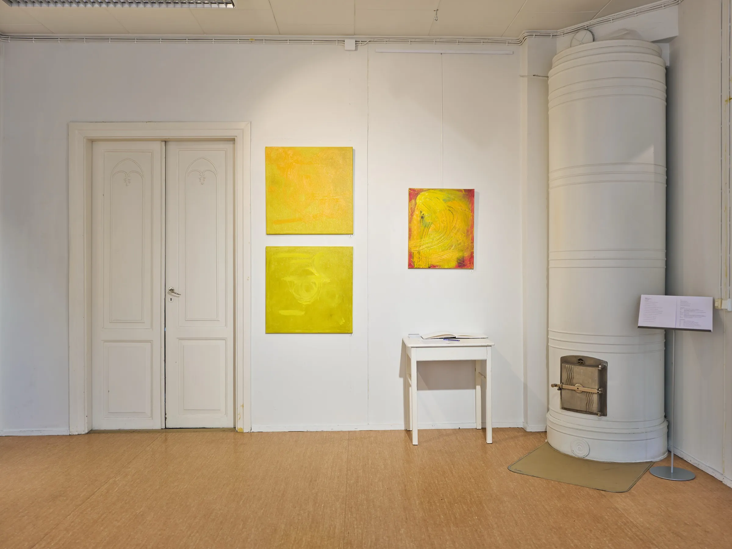 Gallery room in the Kulttuurikonttori with yellow abstract paintings on a white wall and an old tiled stove