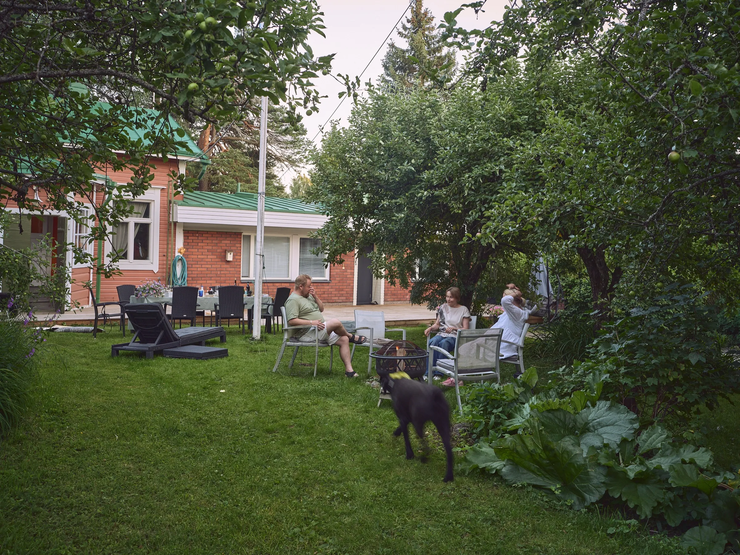 Evening circle of chairs, people and a dog in a garden in front of a brick house