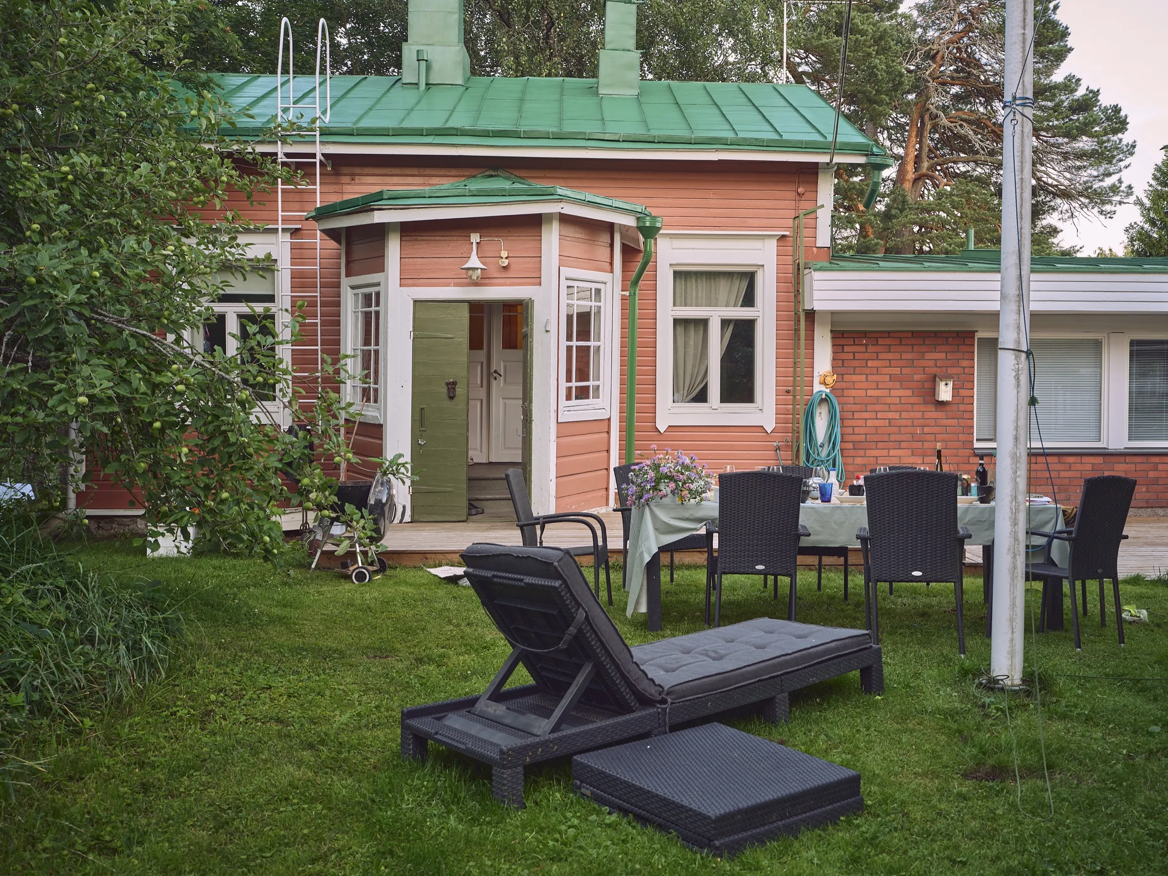 Red house with a green sheet metal roof and a bay window, garden loungers and a set table in front