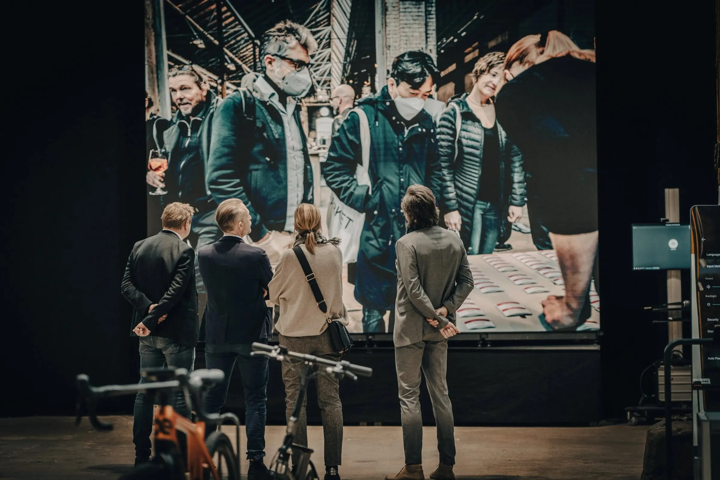 Vier Personen vor Großprojektion in dunkler Halle, Bühne mit Bildmotiv von Menschen, links Fahrräder