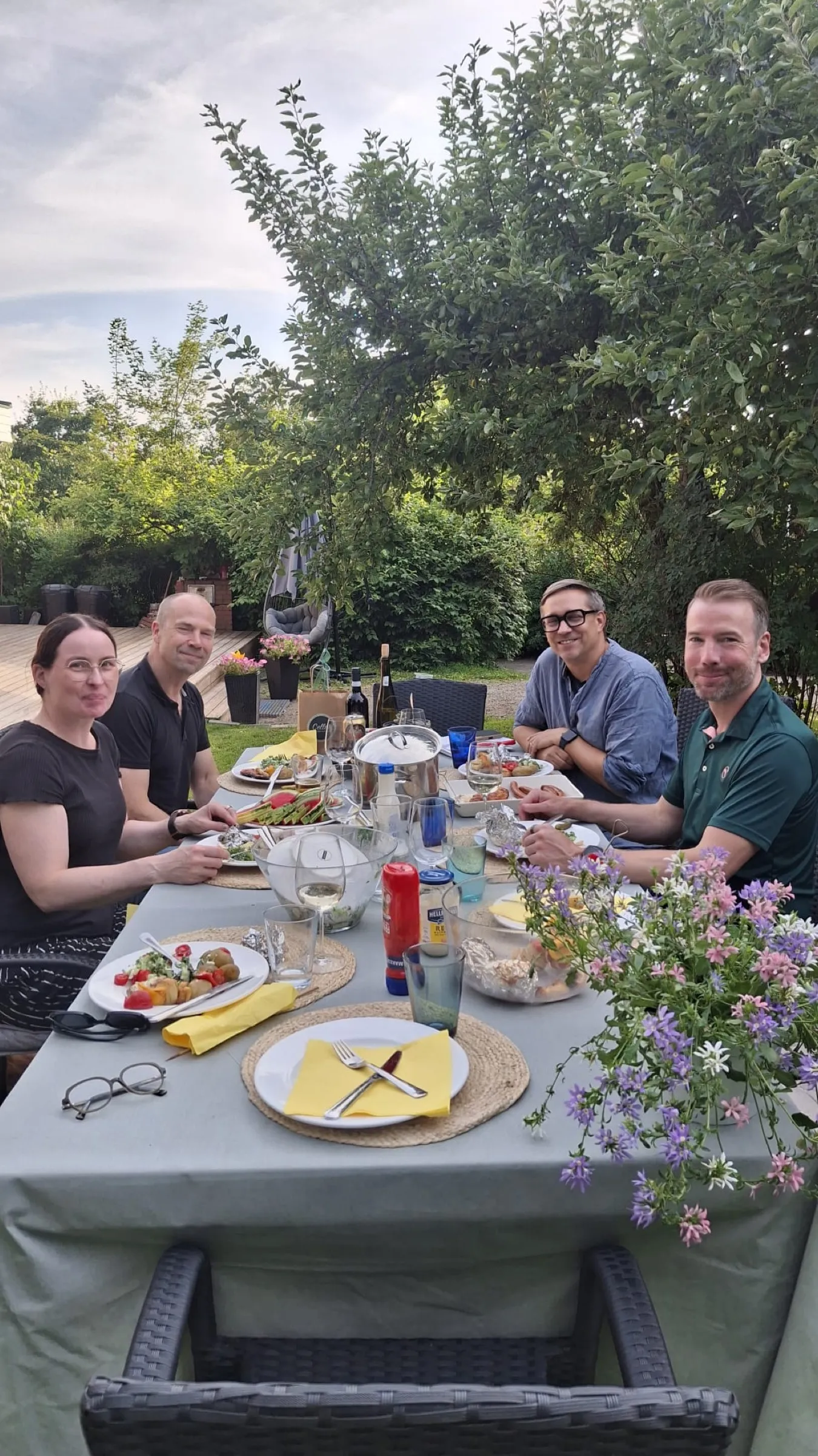 Summer table in the garden, four people eating, flowers in the foreground, portrait format