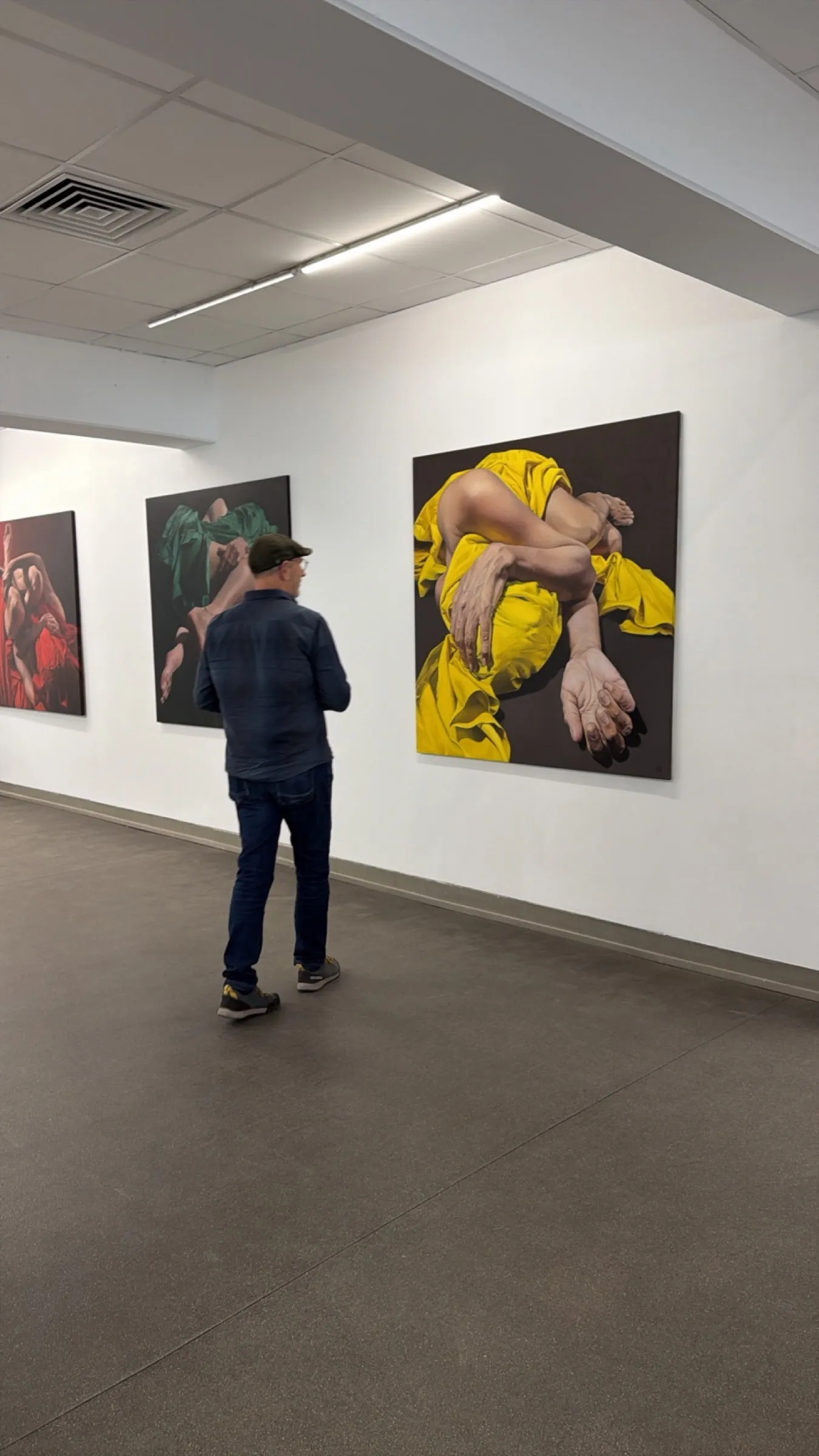 Roman Thomas seen from behind in a white exhibition room in front of three paintings by Bego Riba with bodies in red, green and yellow