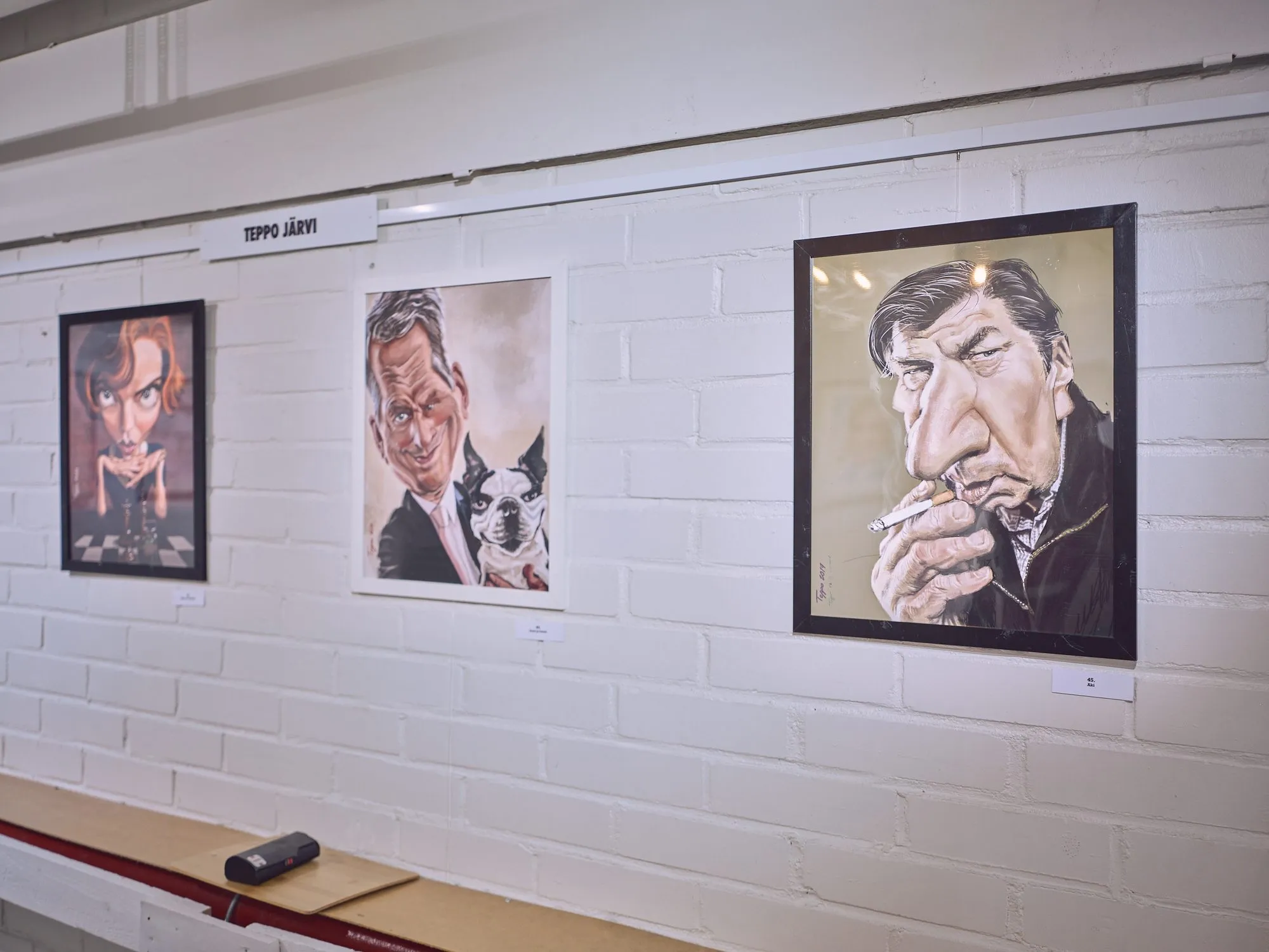 Three caricatures by Teppo Järvi on a white brick wall, in the middle Niinistö with a small dog