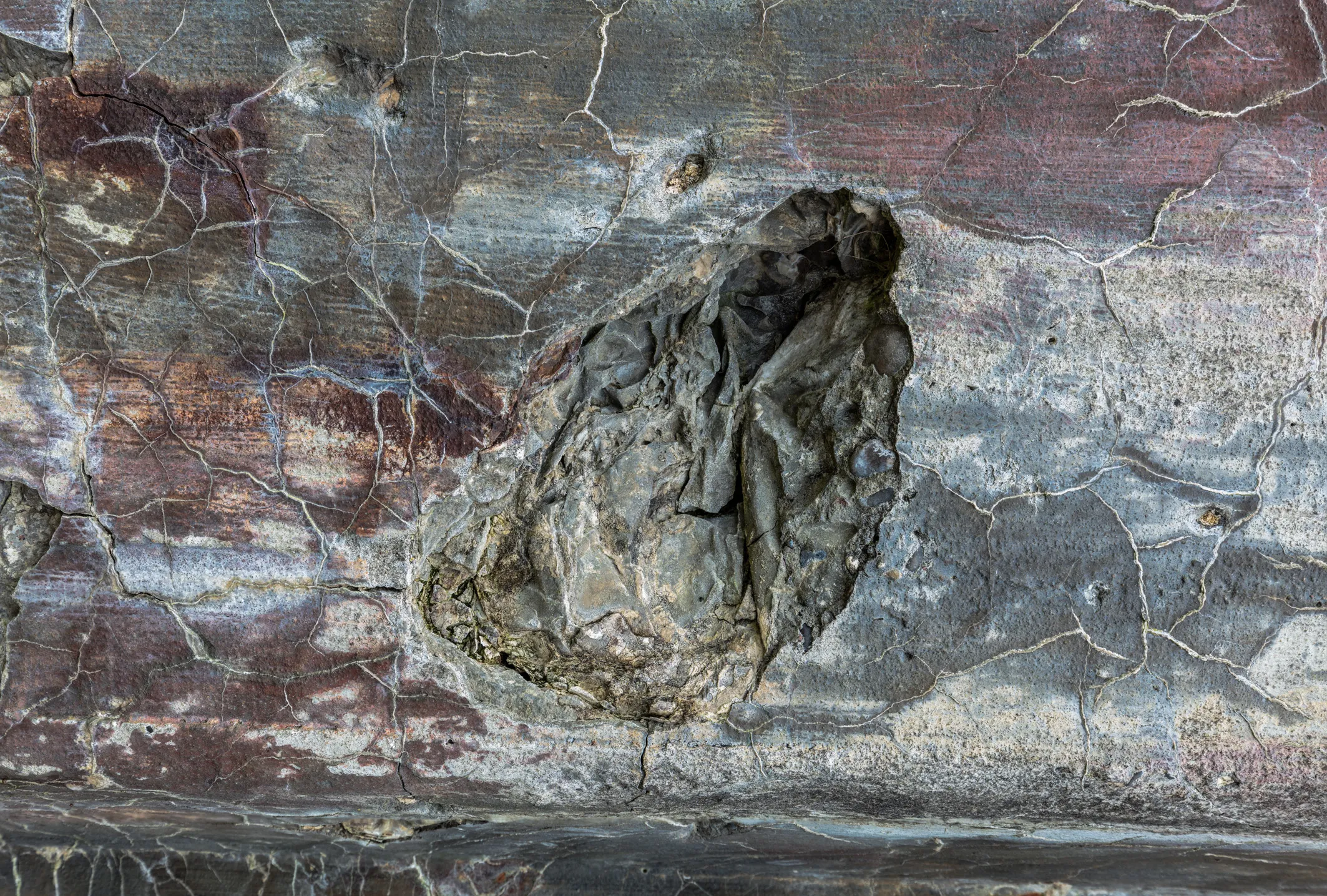 Aufgebrochene Betonwand mit grauem Auswuchs in der Mitte, rotbraune und graue Verfärbungen, feine Risse