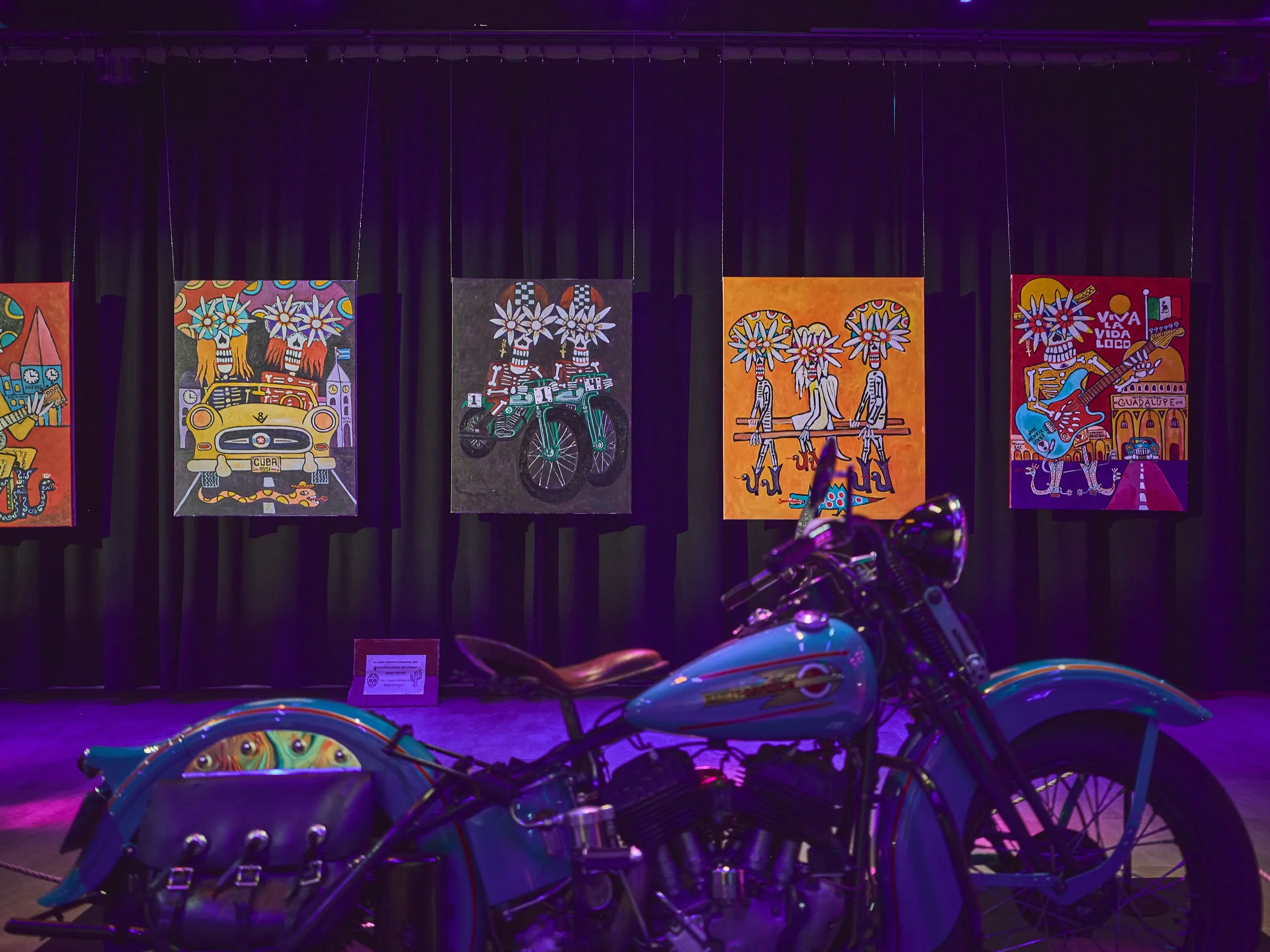 Motorcycle with a blue tank in front of a row of colourful paintings