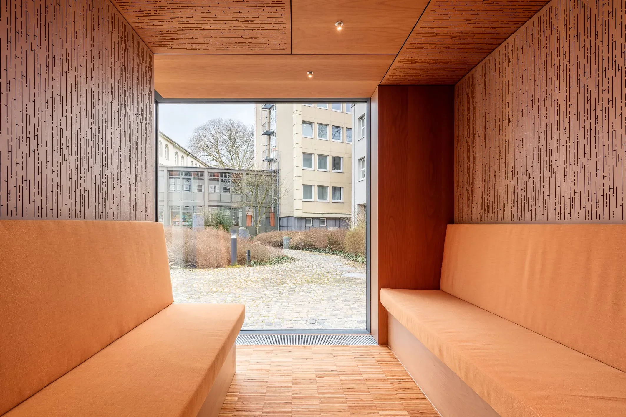Innenraum mit Sitznische aus Holz, Fensterfront zum gepflasterten Innenhof, dahinter Wohnbauten bei Tageslicht