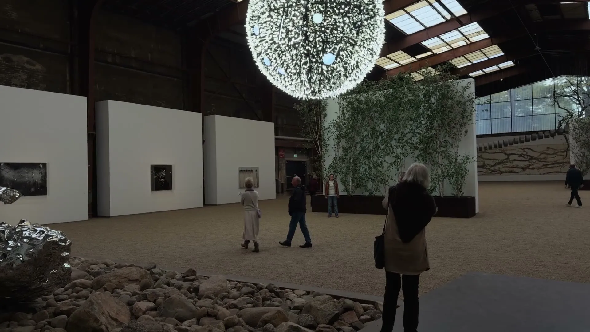 A slow camera move through the great hall, the glowing sphere above, visitors on sand and stones below