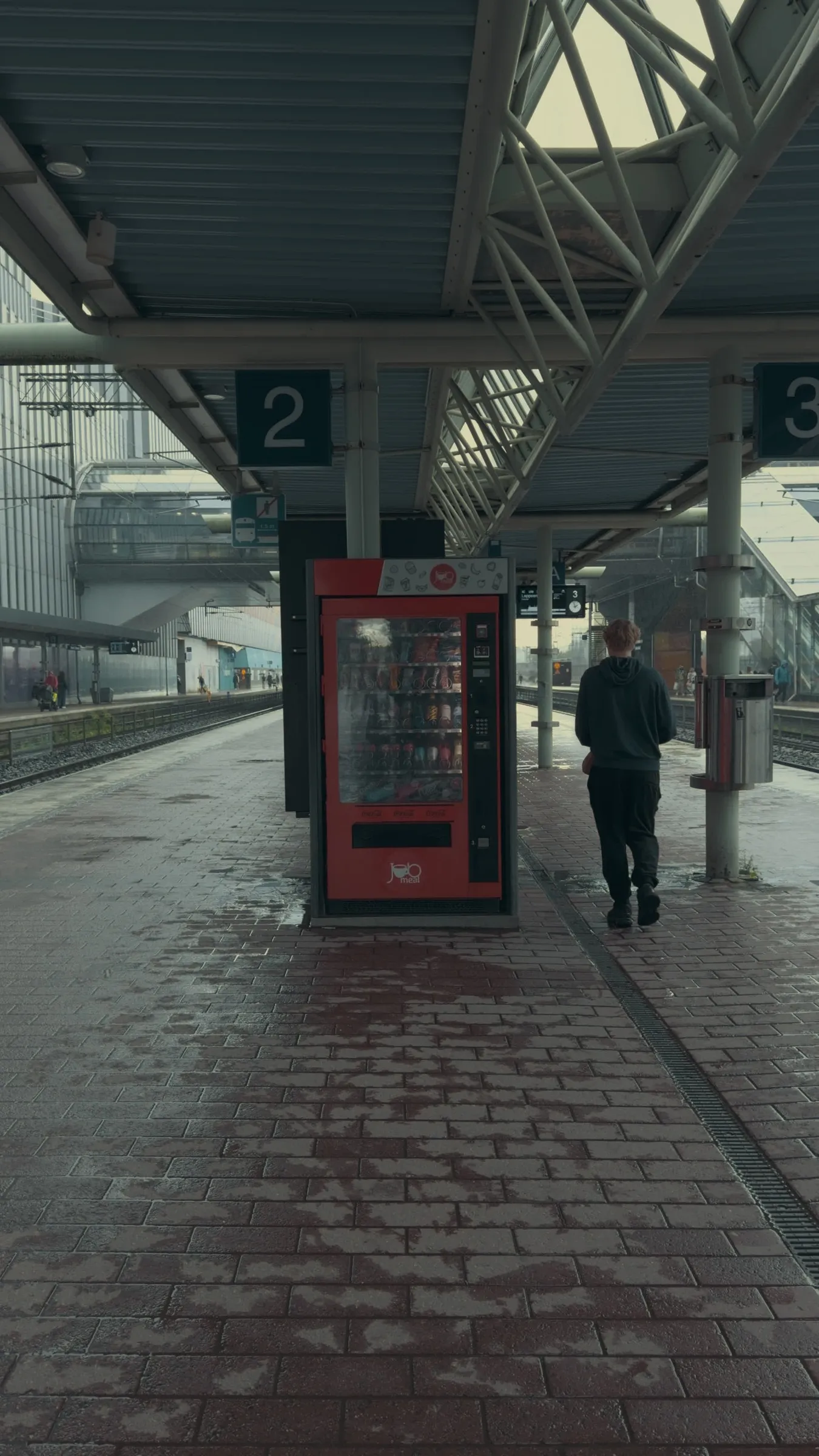 Rainy platform with a red vending machine, one person waiting