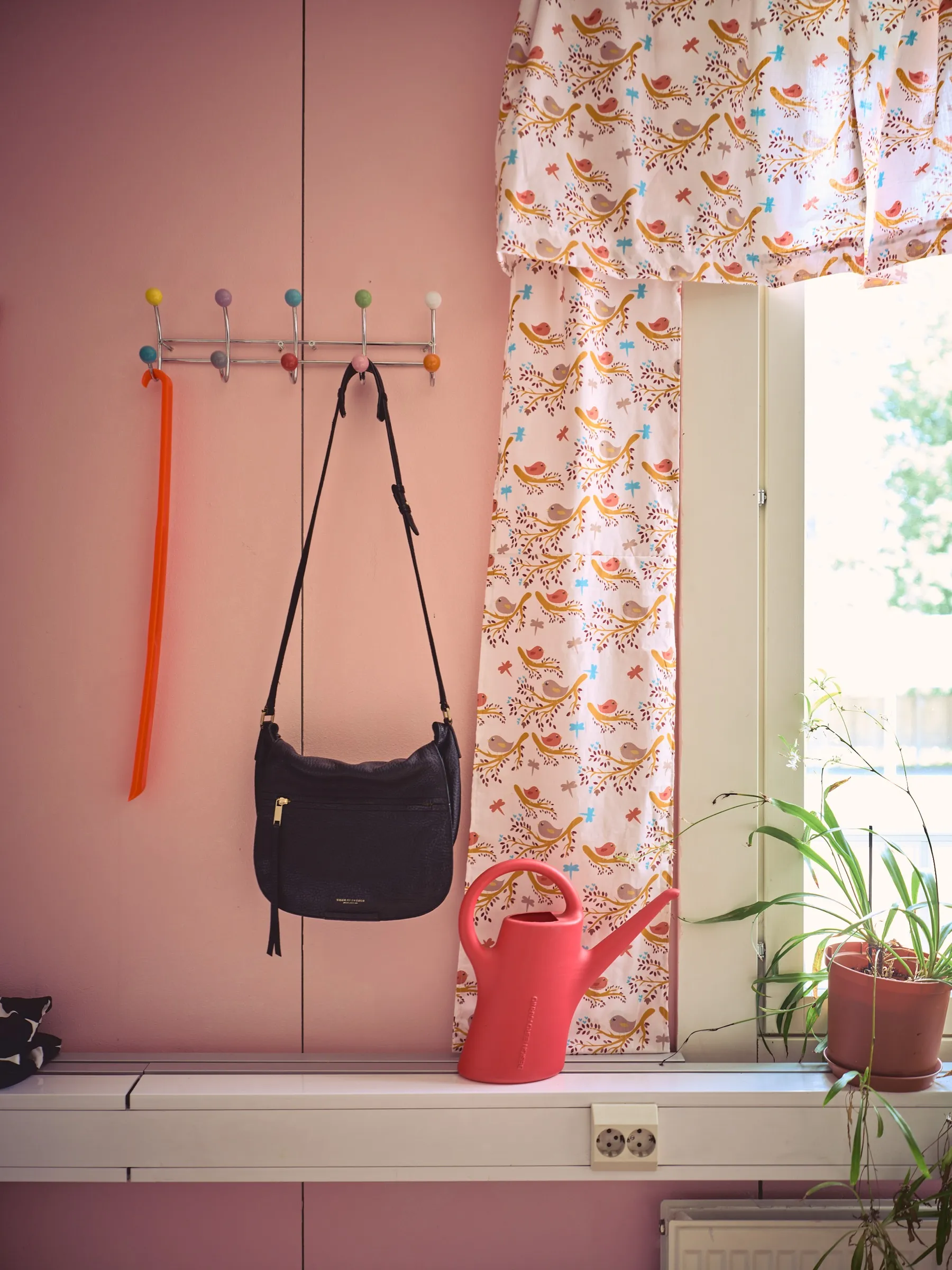 Pink wall with a colourful hook rail, a black handbag and a red watering can by the window