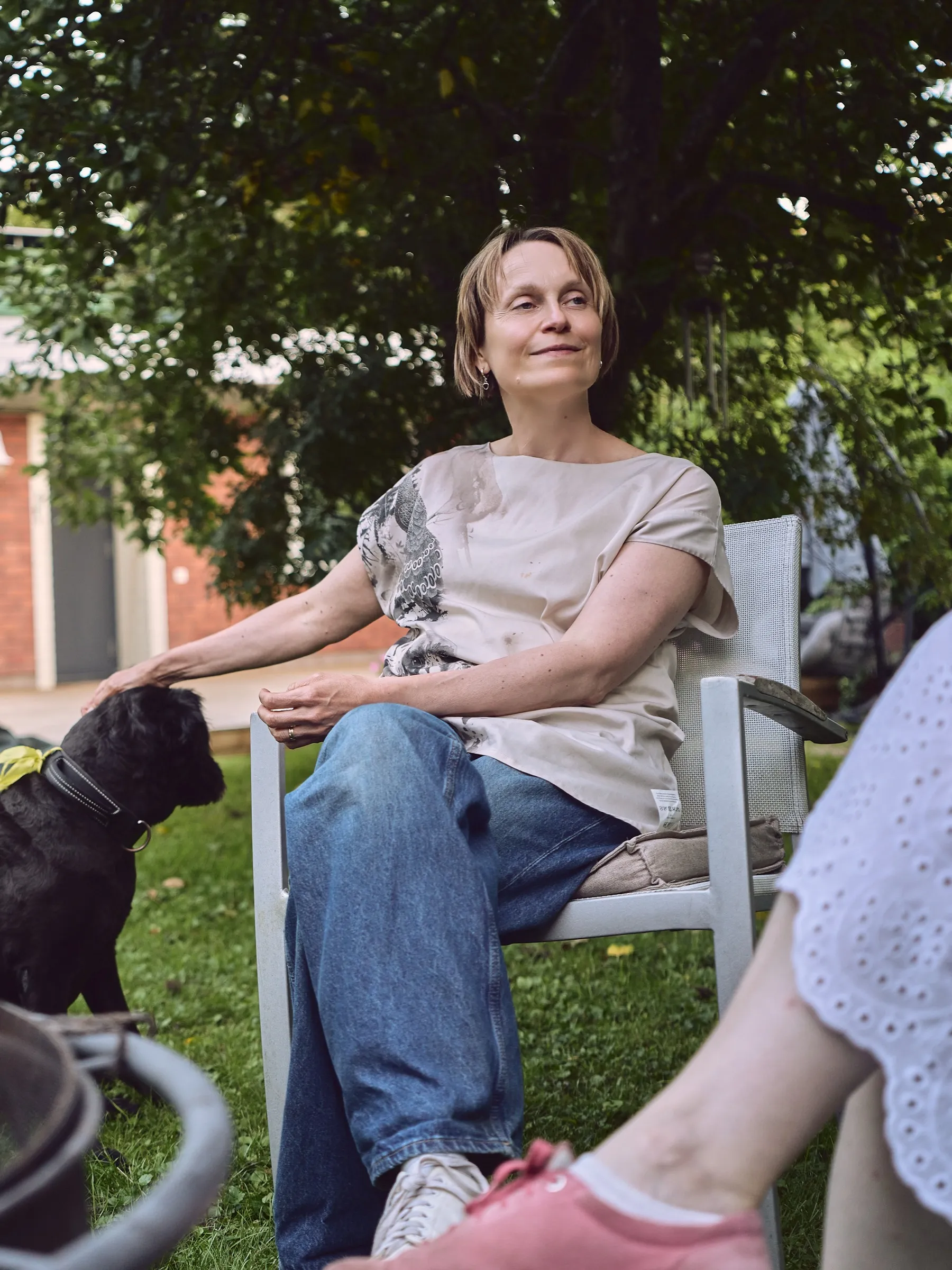 Woman sitting relaxed on a garden chair under trees, a black dog beside her, portrait format