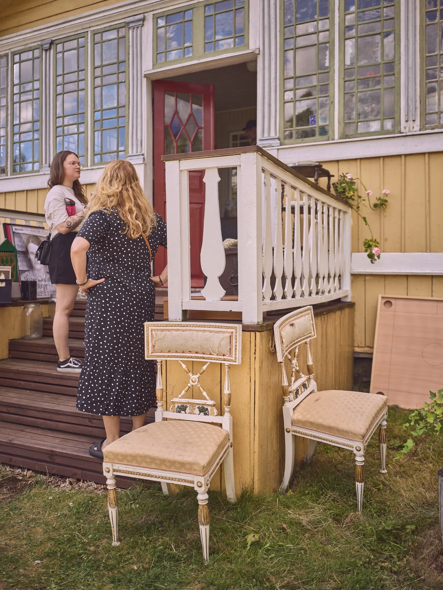 Two women talking on the steps of the yellow house, two old upholstered chairs in front, portrait format