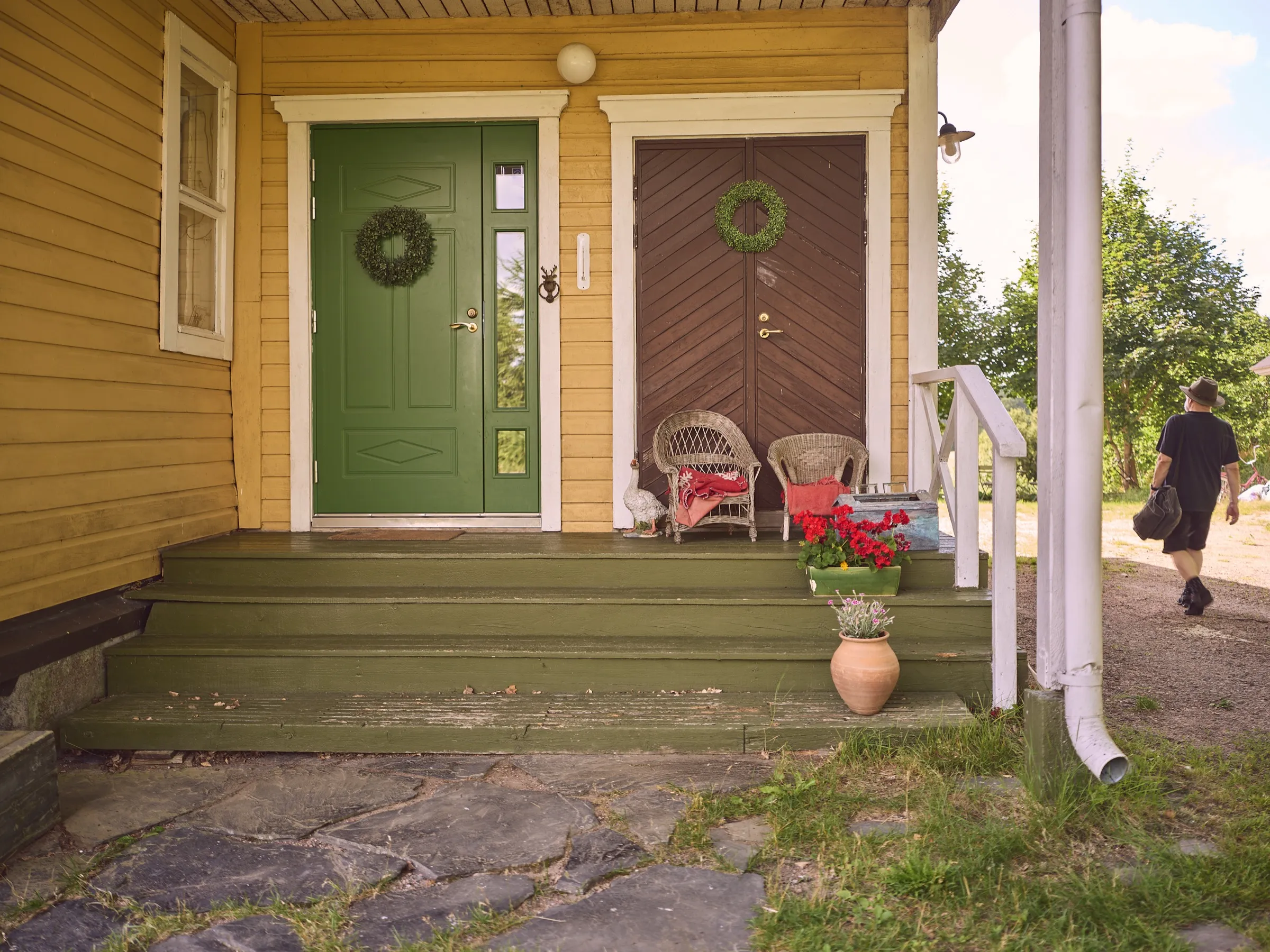 Porch of the yellow house with a green and a brown door, wreaths and wicker chairs