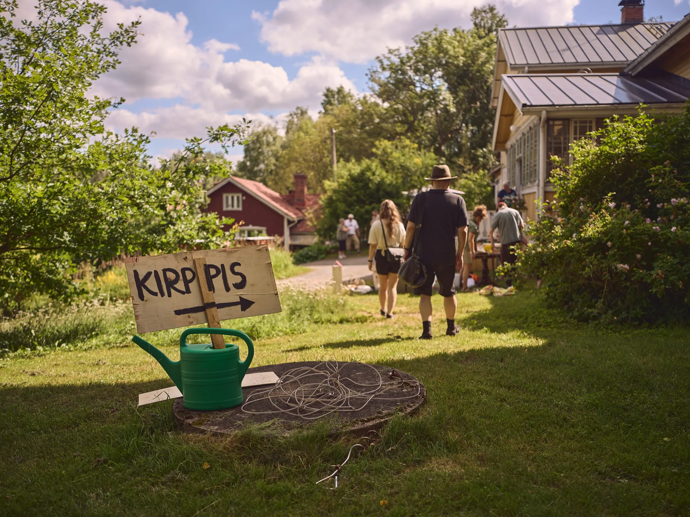 Hand-painted KIRPPIS sign with an arrow on a green watering can, people walking towards the yard behind it