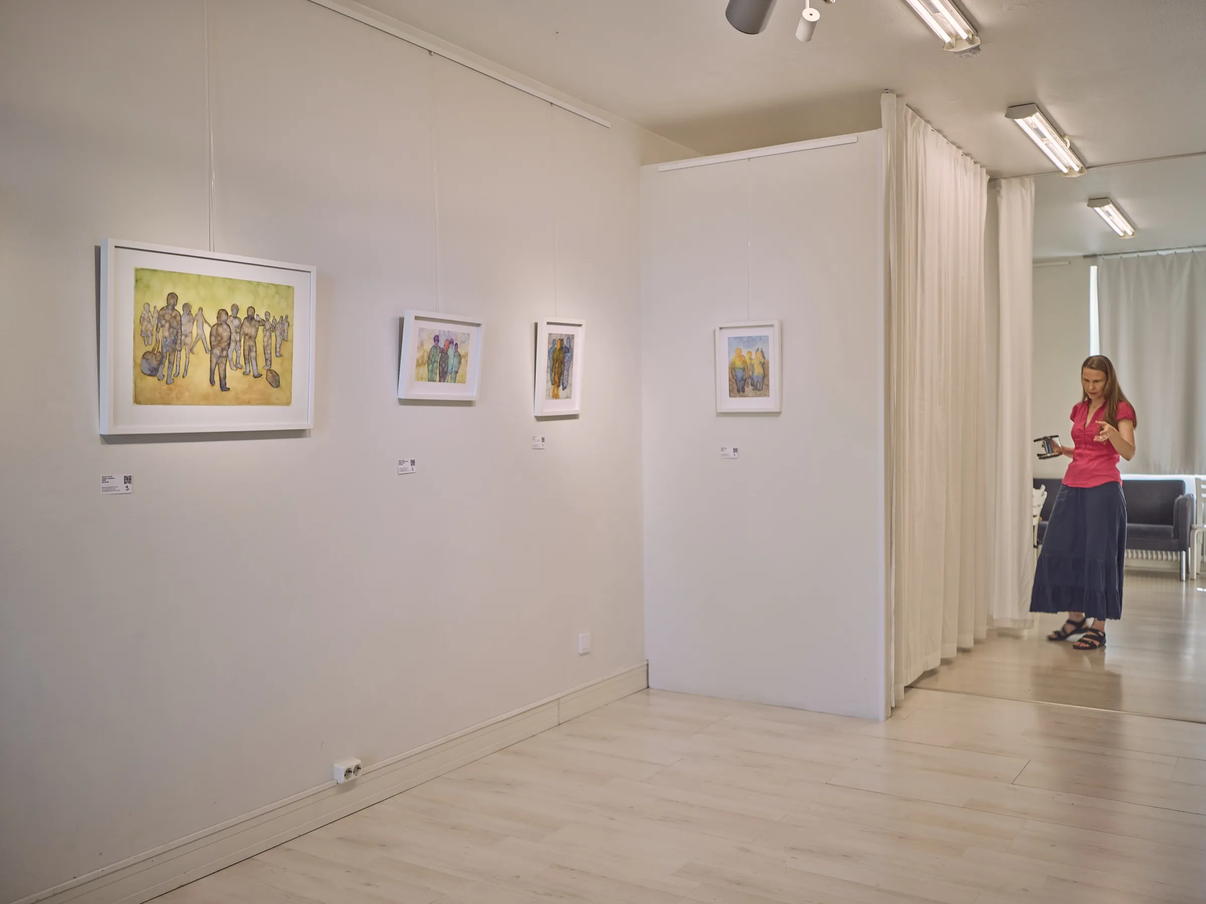 White gallery wall with small watercolours, a woman by the curtain in the background