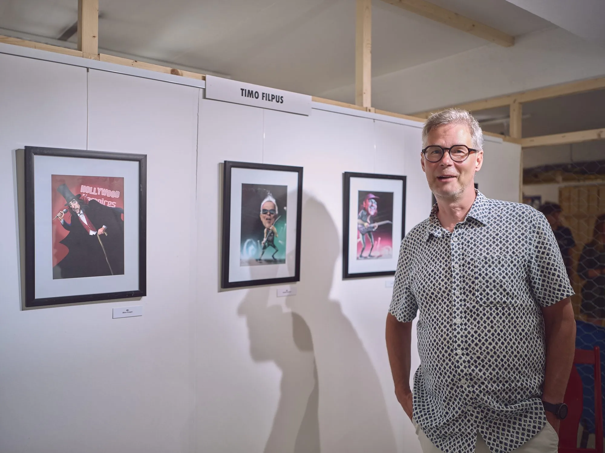 Timo Filpus stands smiling at his exhibition wall with framed musician caricatures