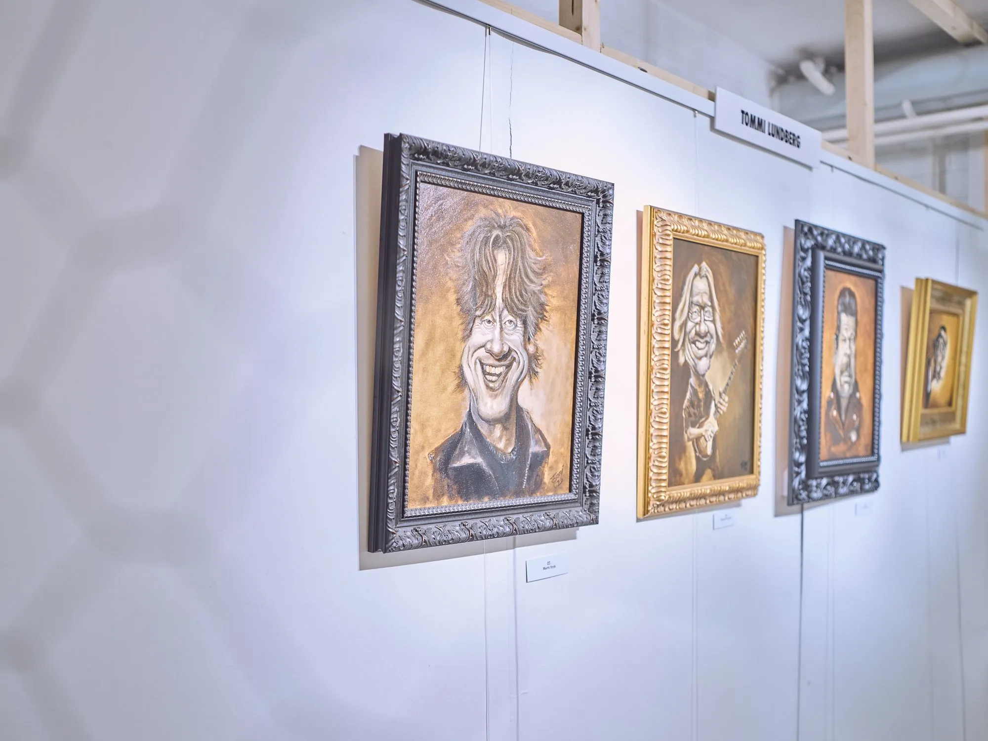 A row of oil caricatures on a white wall under the name plate Tommi Lundberg