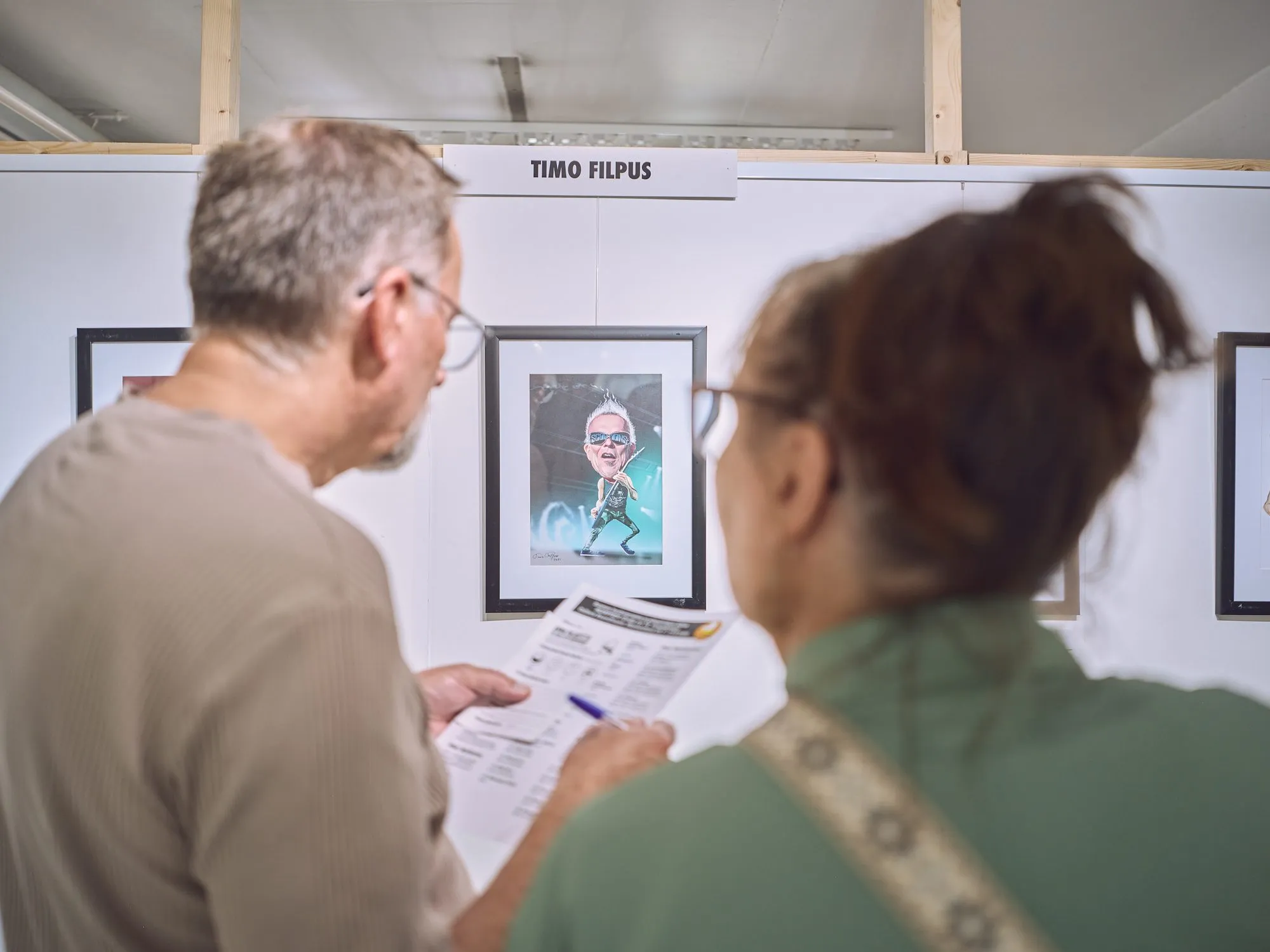 Two visitors look at a musician caricature, one holding the work list