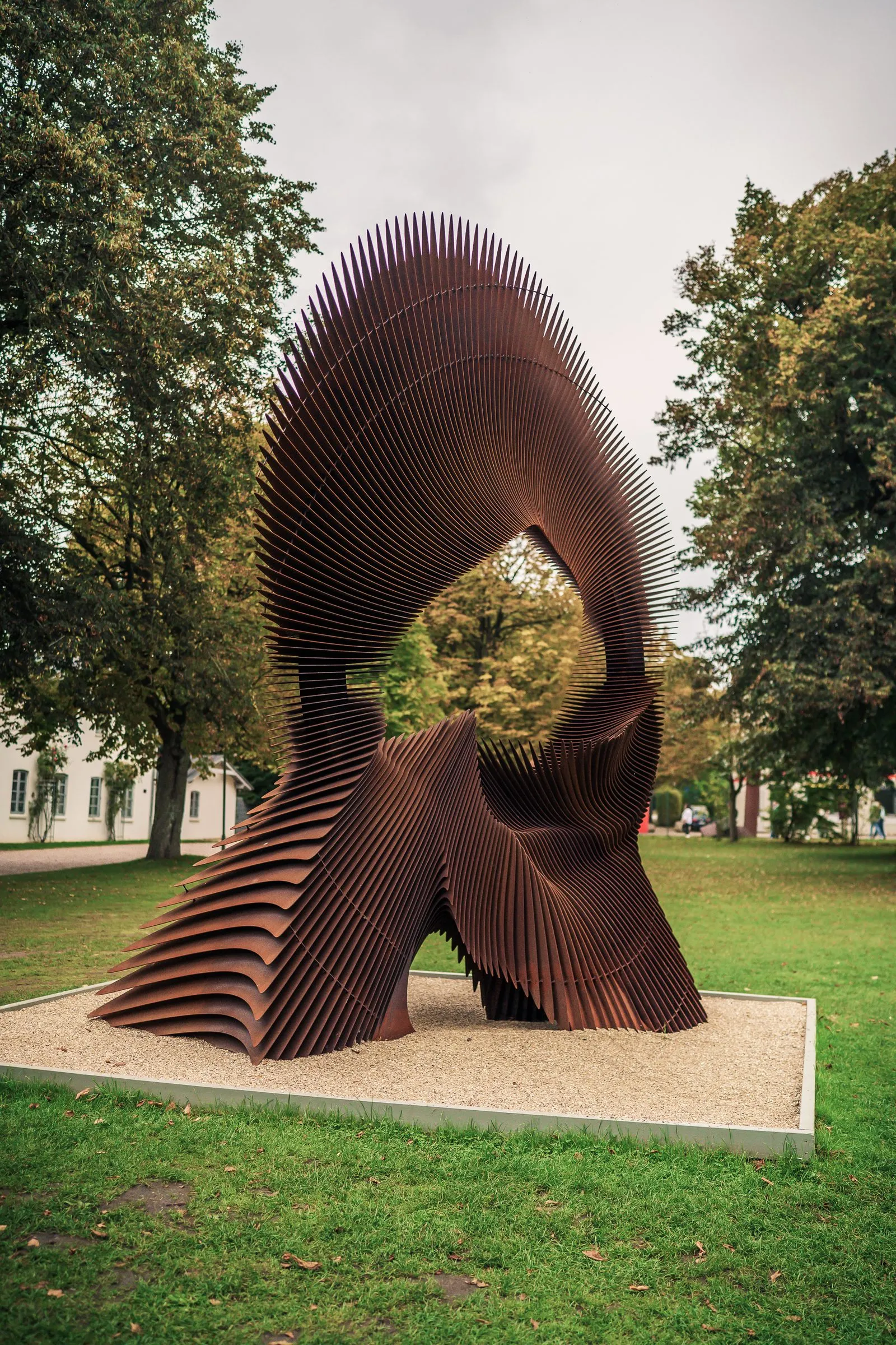 A tall sculpture of many curved, rust-brown slats on a pale plinth in the park