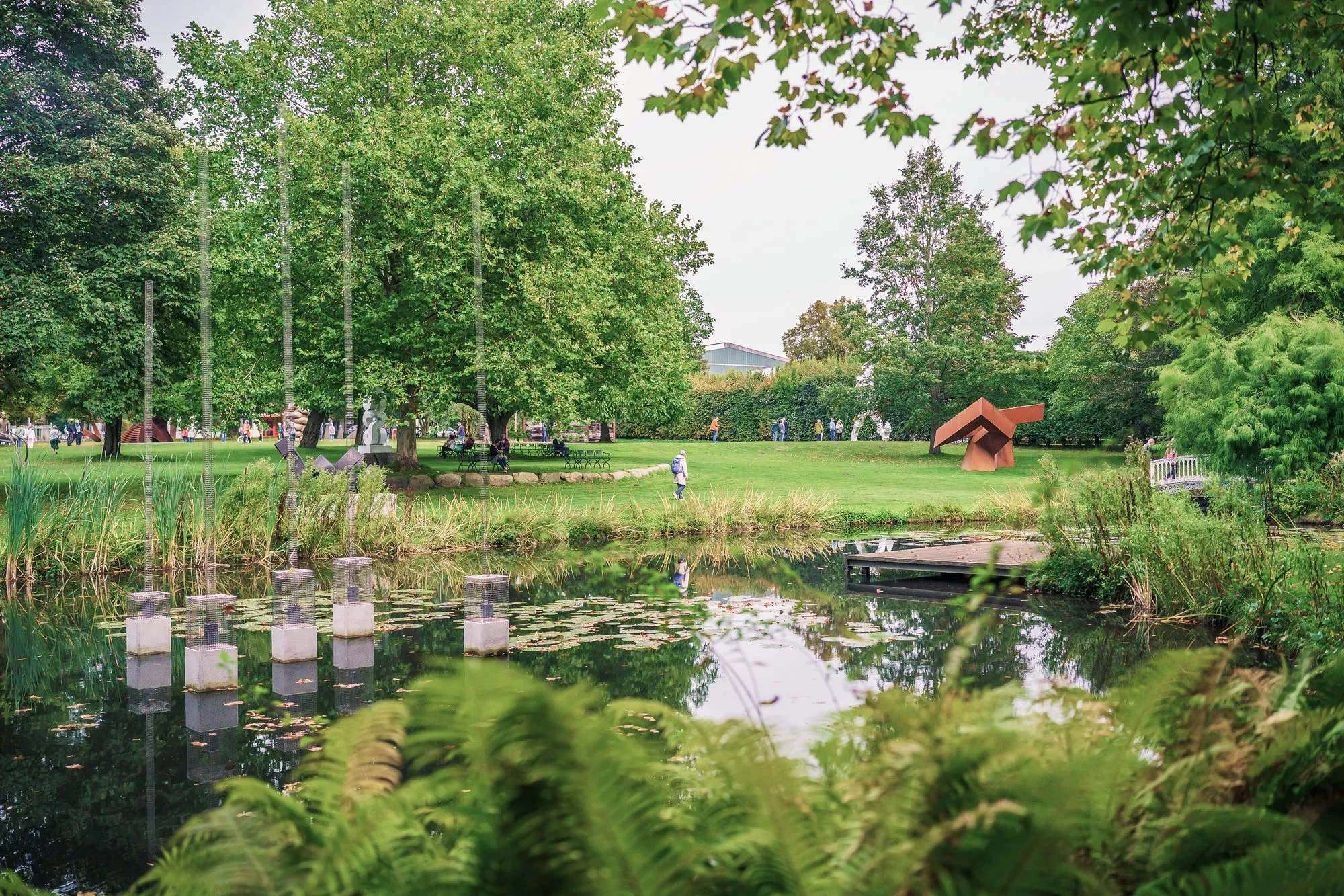 A pond with water lilies and pale steles in the water, lawn, trees and a red sculpture behind
