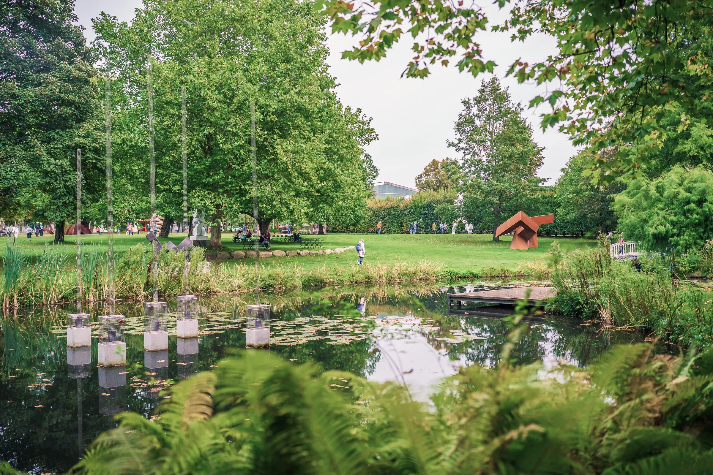 A pond with water lilies and pale steles in the water, lawn, trees and a red sculpture behind