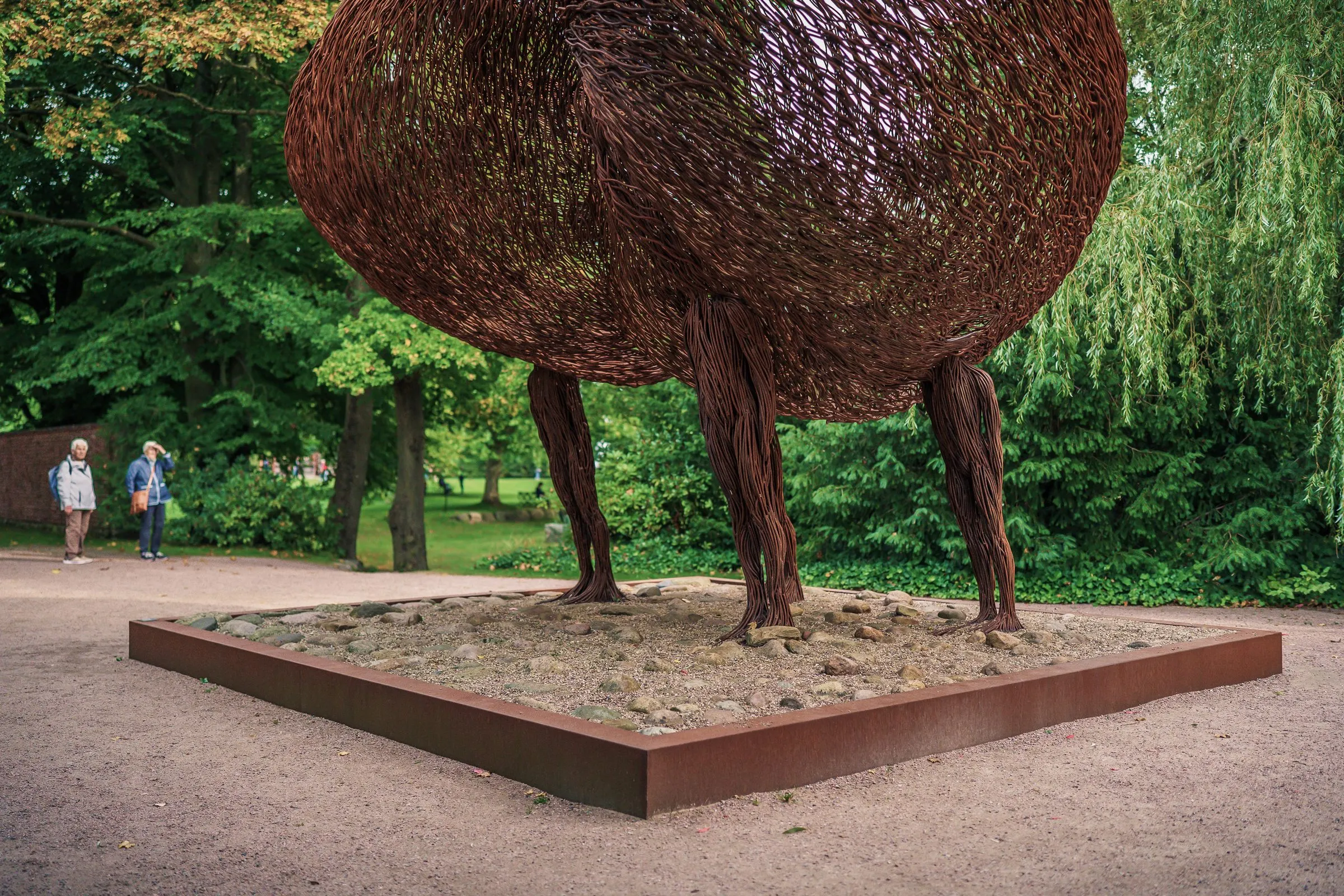 A huge oval body of steel wire on four thin legs in a rusty frame, visitors in the background