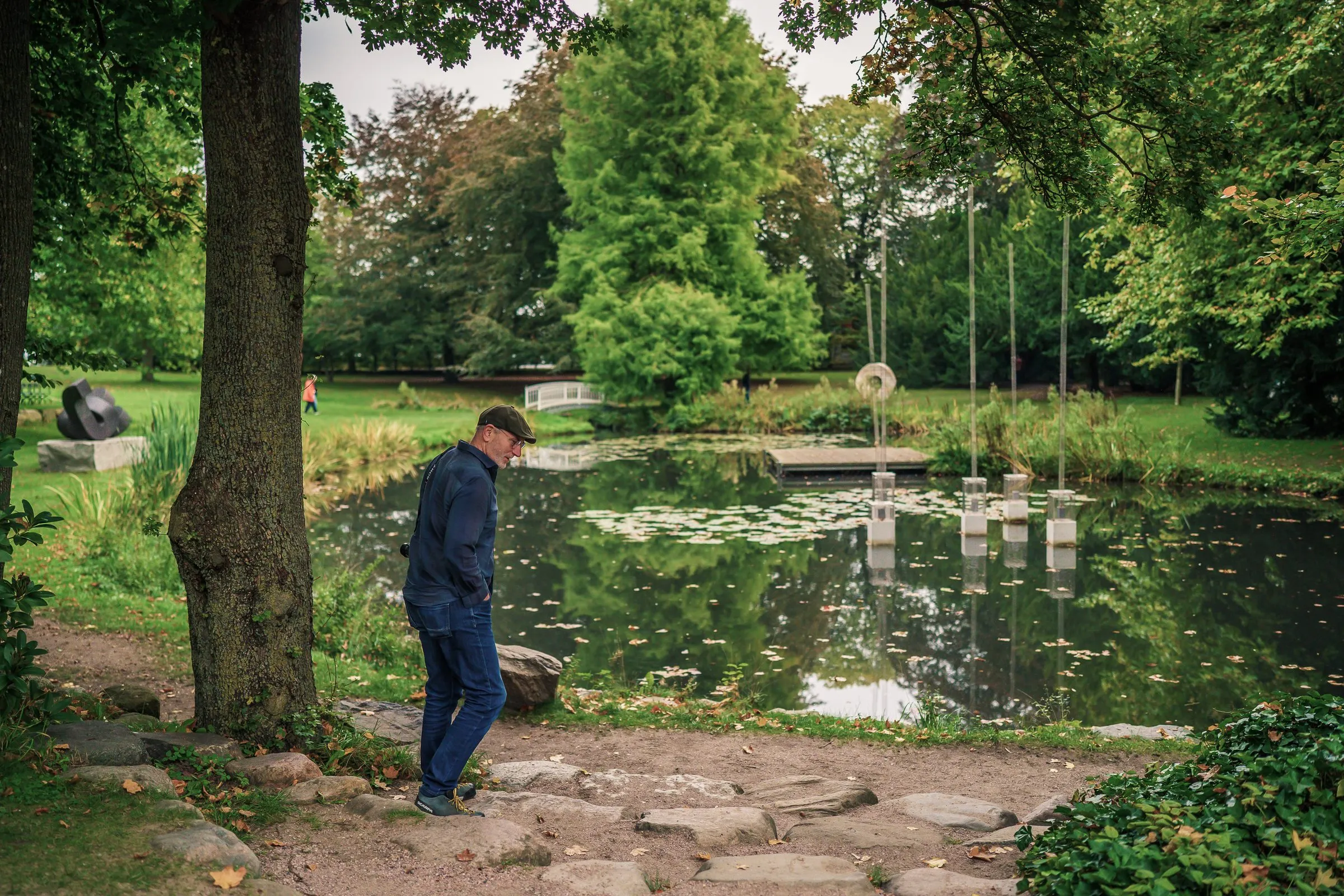 A man walks over stepping stones by the pond, sculptures on the bank behind