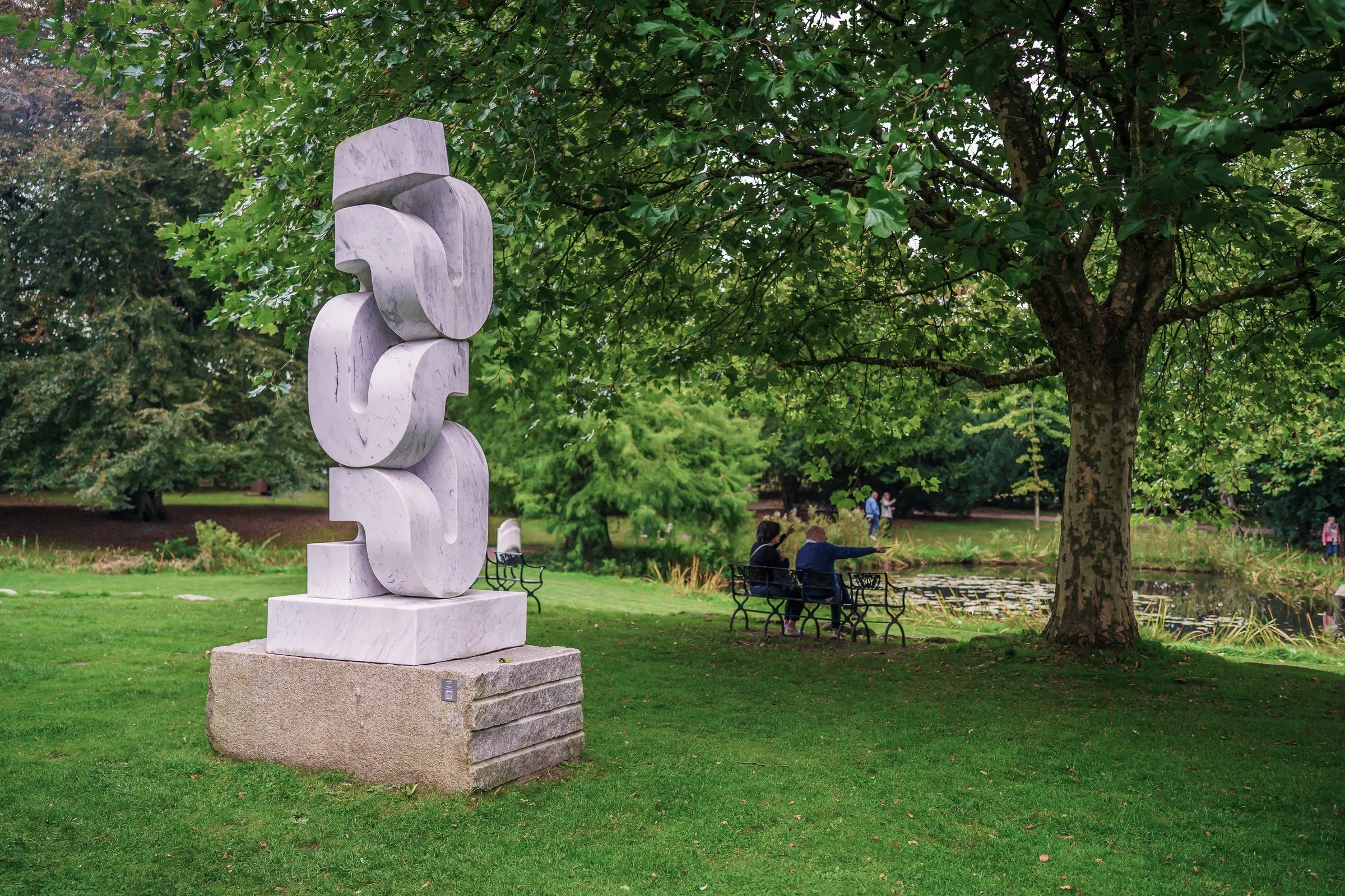 A white stacked stone sculpture on a plinth, visitors on a bench under trees behind it