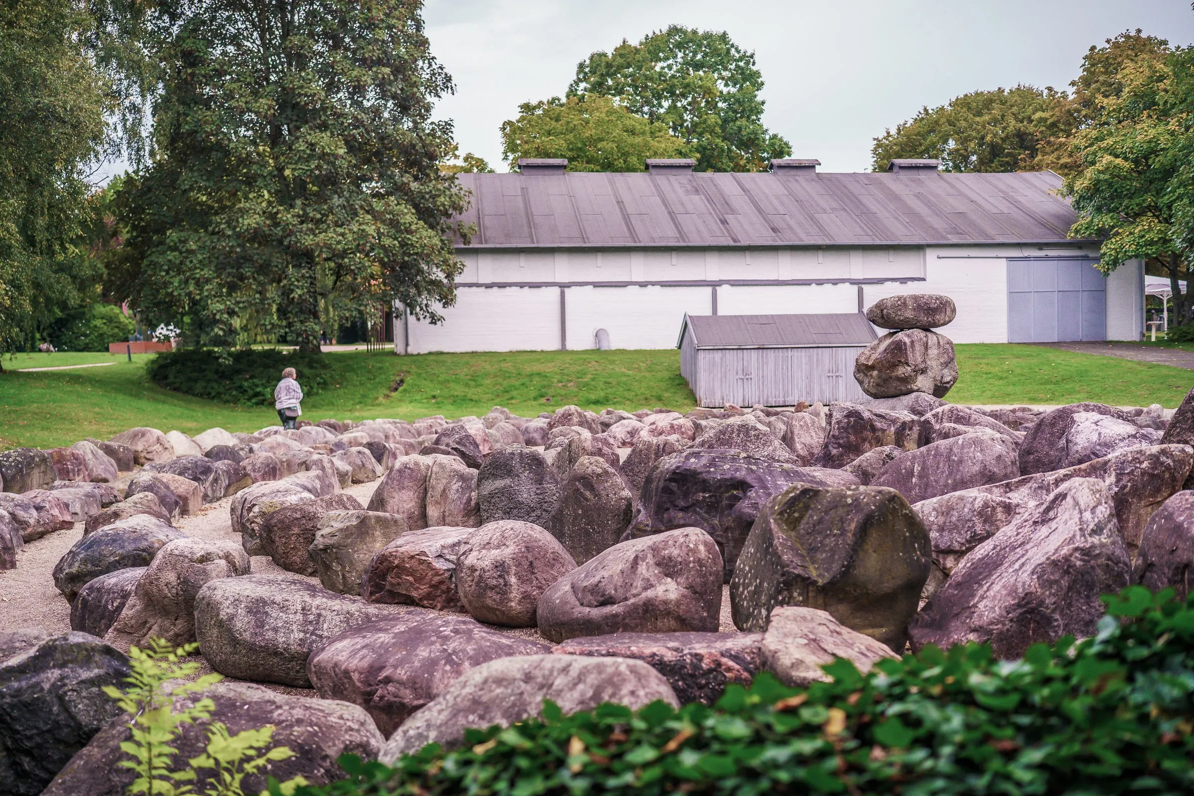 A field of many large boulders, a tower of stacked stones on one of them, a long white hall behind