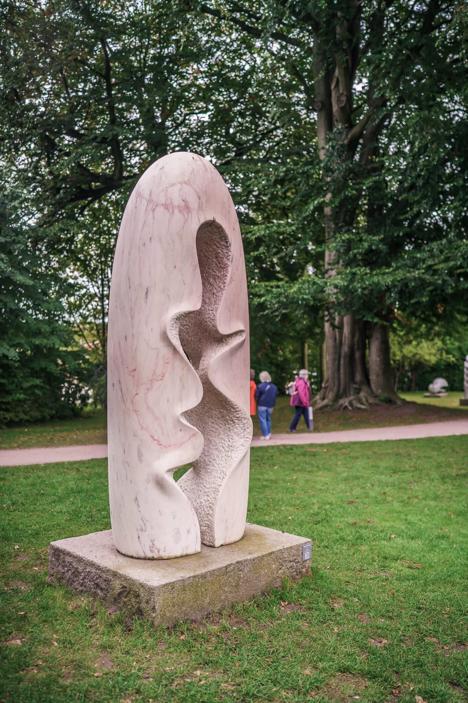 A tall white stone form with cut openings on a plinth, old trees behind