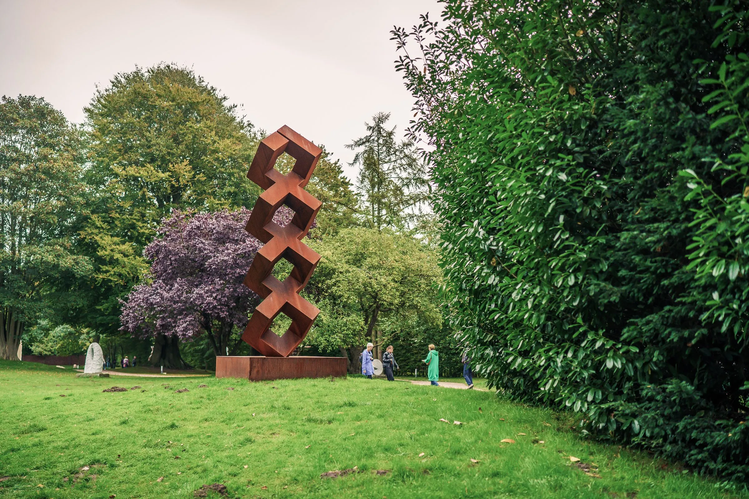 A tower of rust-red hexagonal frames on the lawn, a hedge and a tree with dark red leaves beside it