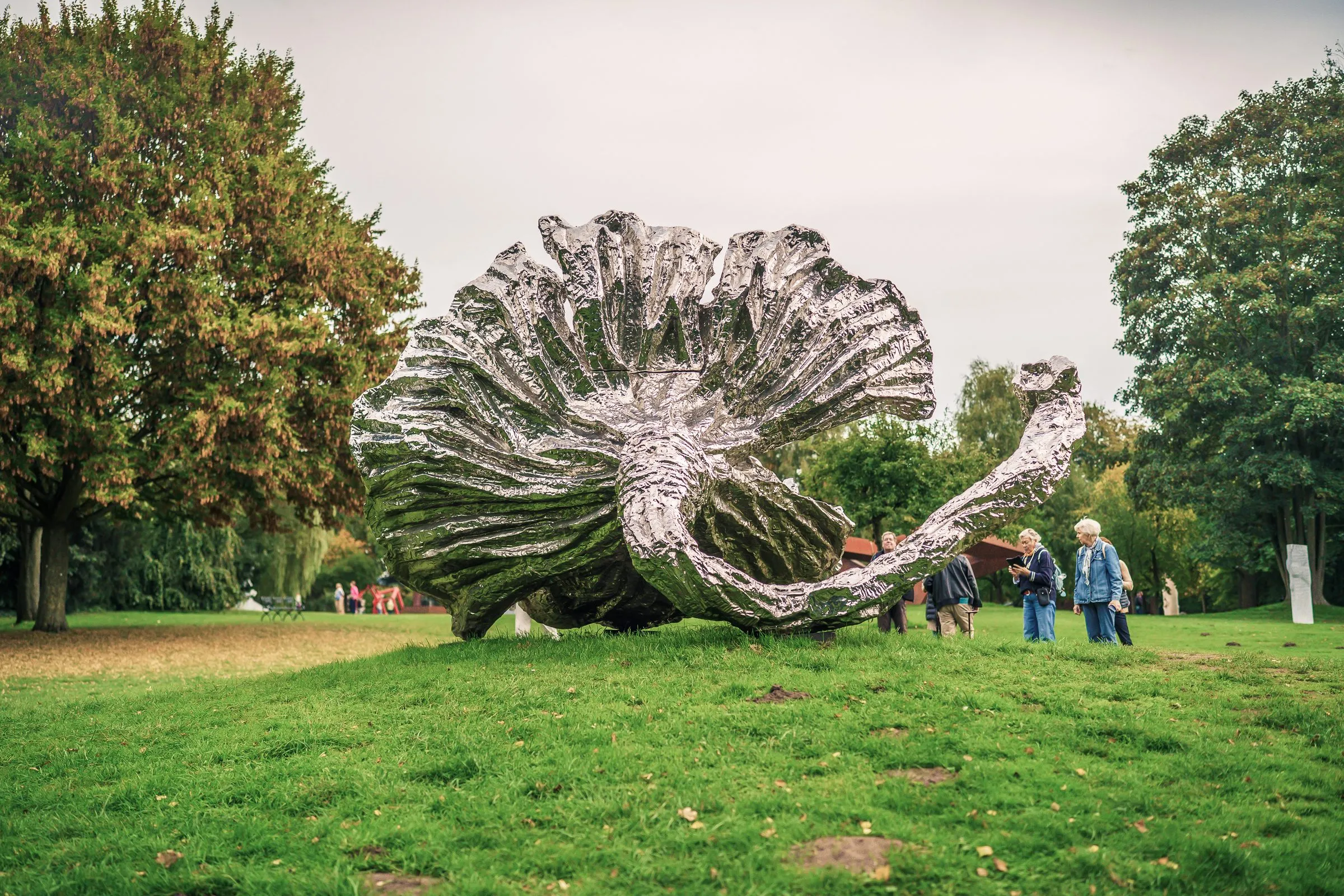 A large, shiny silver, crumpled form like a shell on the lawn, visitors behind it