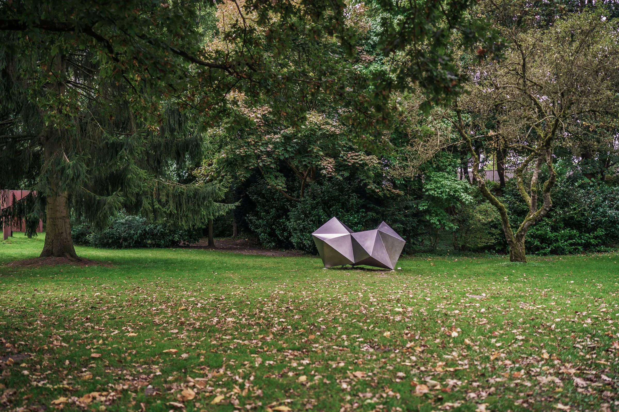 A small, angular stainless steel sculpture on a wide lawn with autumn leaves