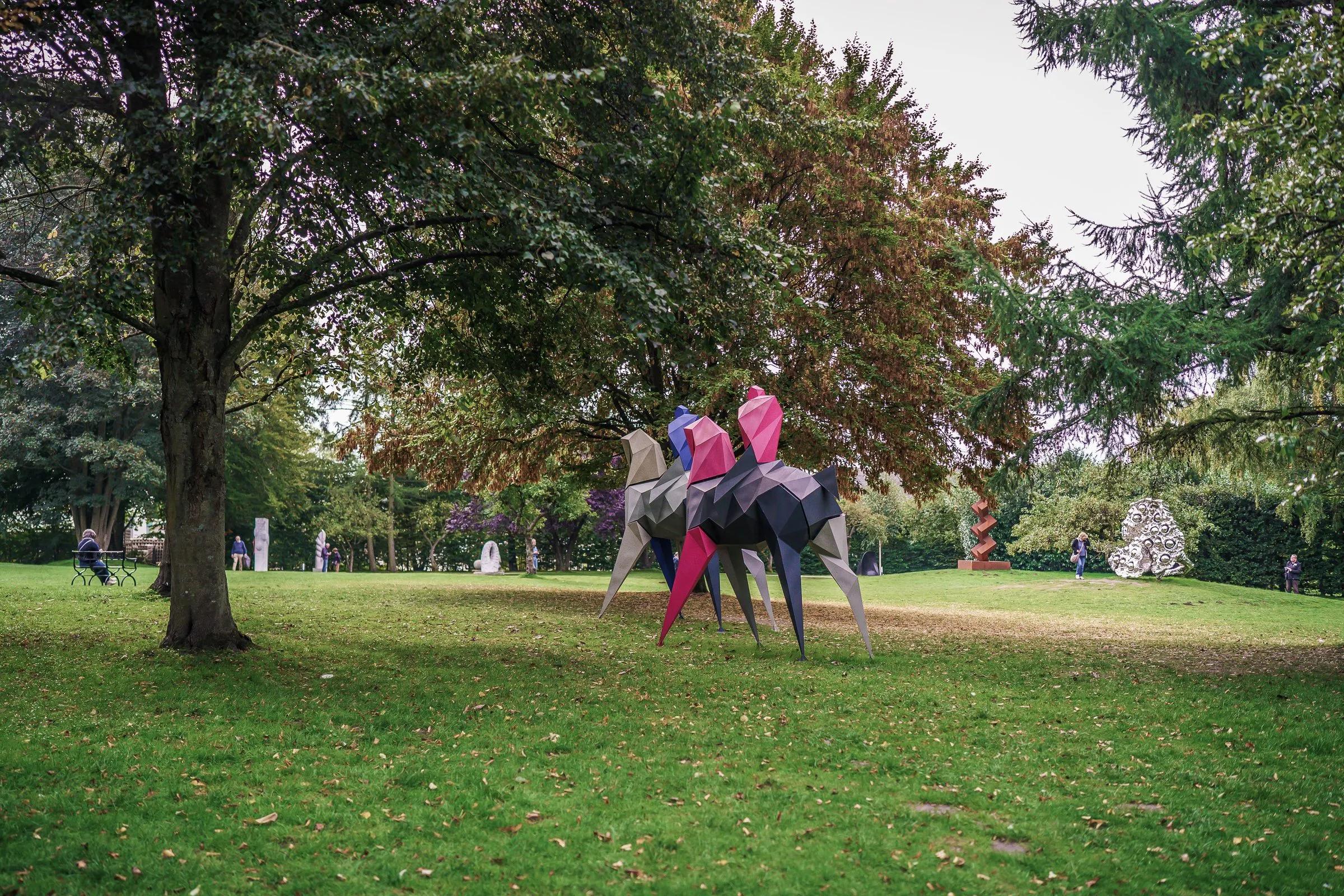 A colourful faceted horse sculpture with a pink figure on the lawn under trees
