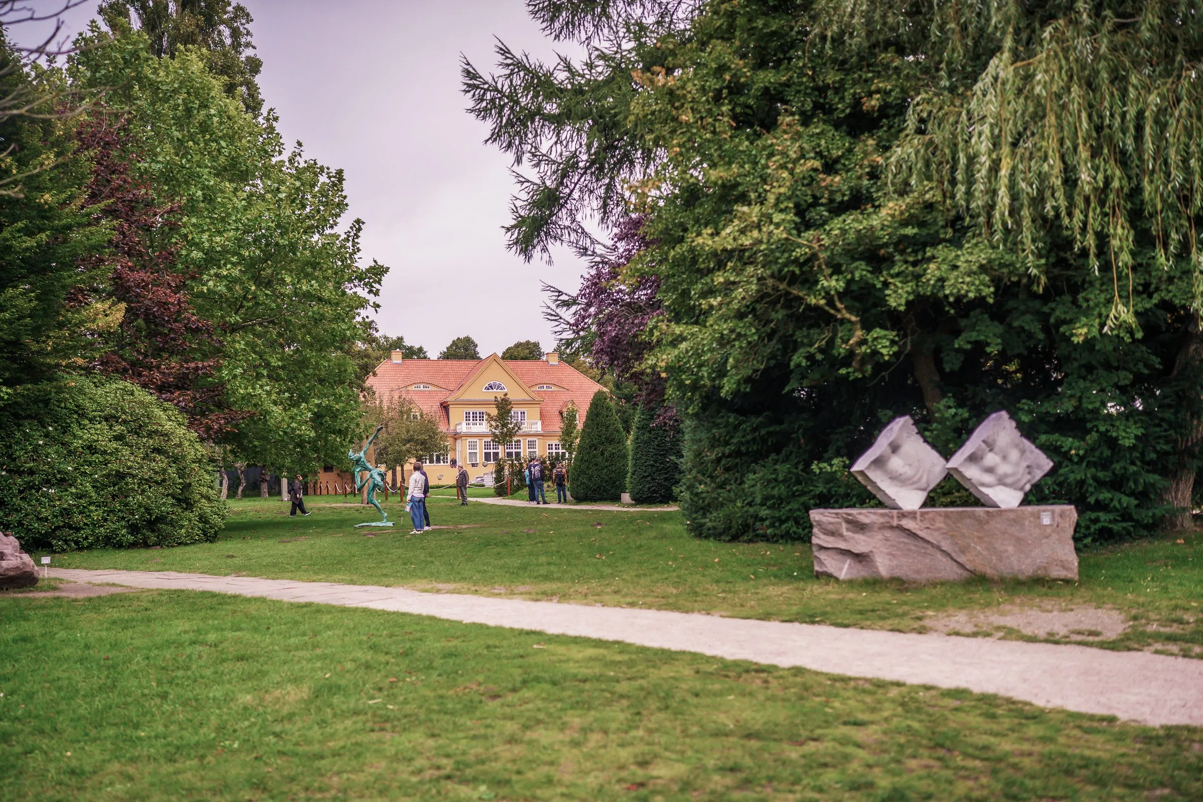 A path through the park with a stone sculpture of two diamonds, a red brick house in the background