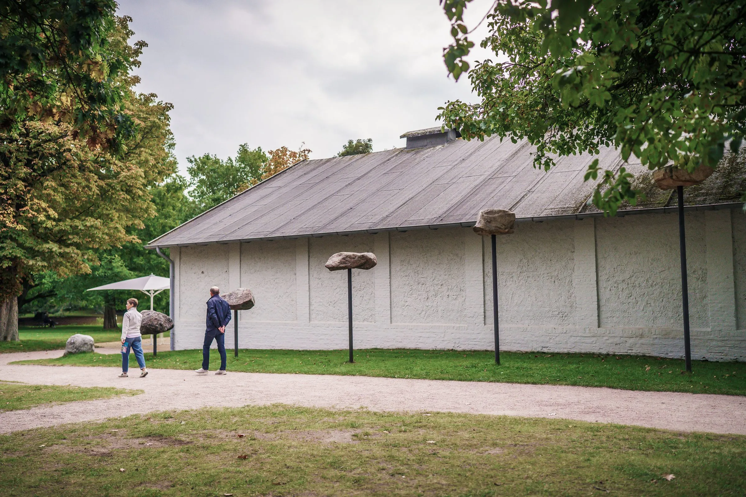 A long white-plastered hall with a grey roof, stones on poles and visitors on the path in front
