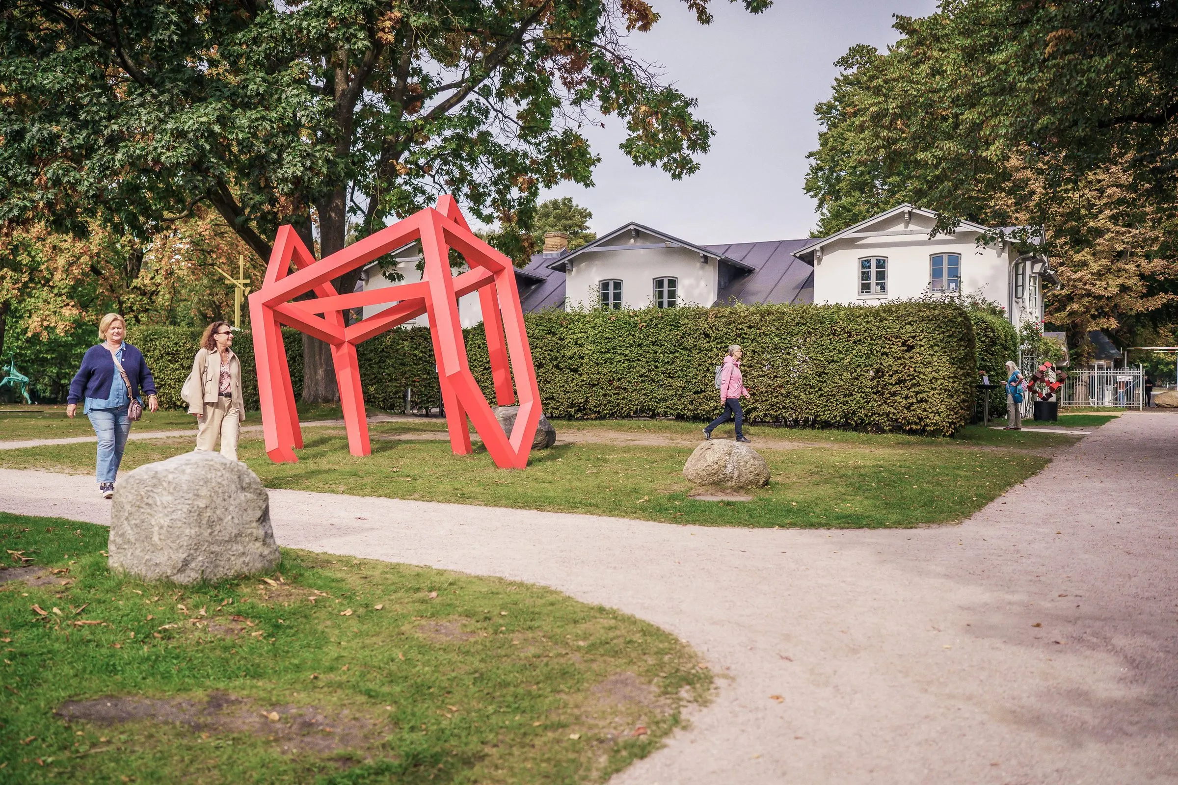 A red sculpture of open, angular frames beside a curving path, visitors next to it