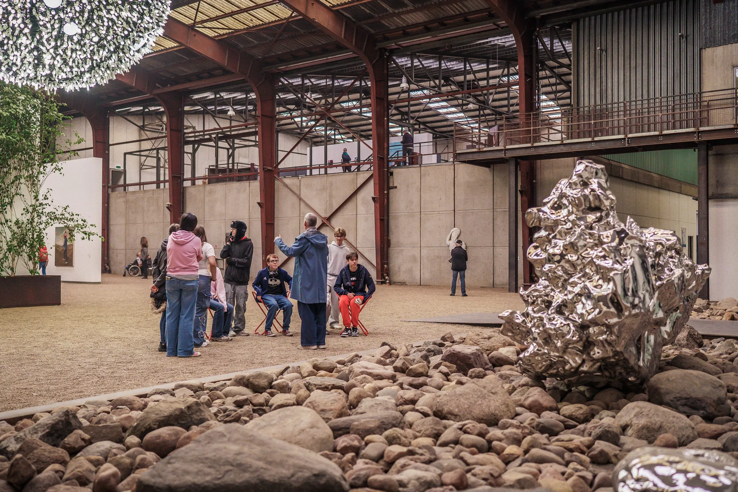 An industrial hall with a gravel floor and stones, a shiny silver sculpture, visitors and a large light object under the roof