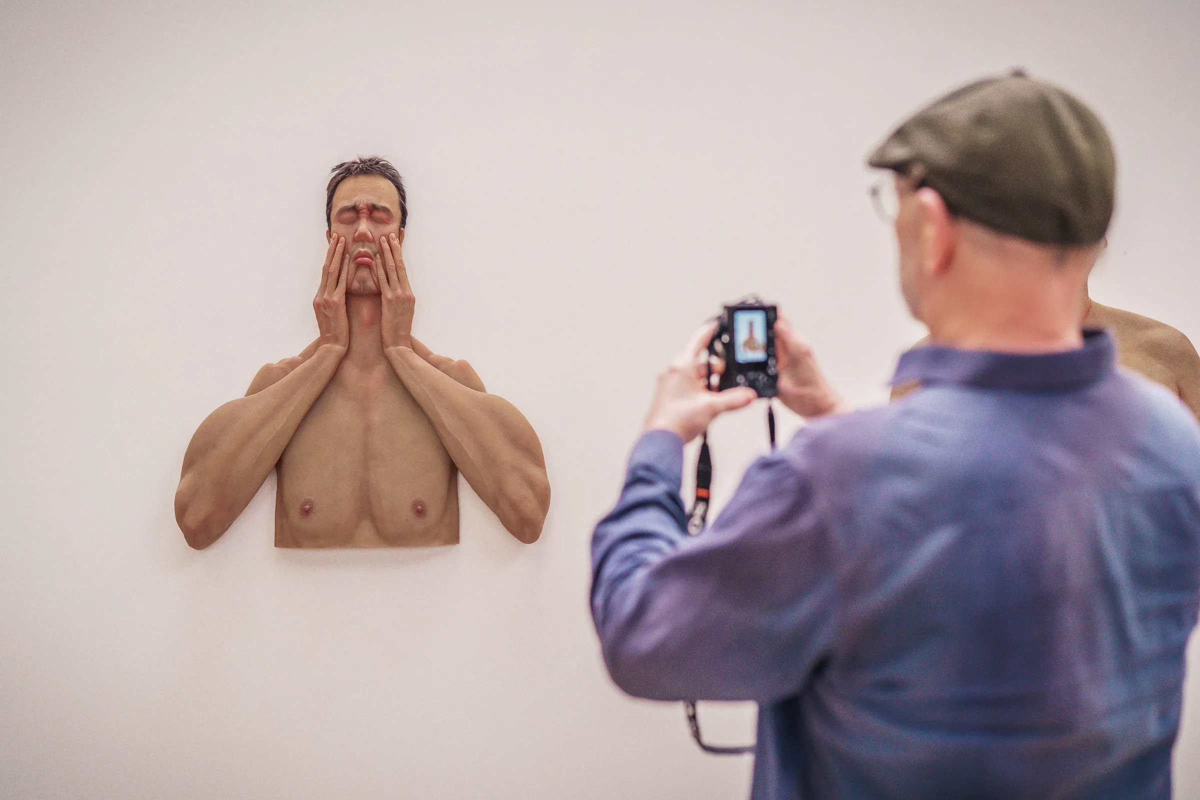 A man photographs a wall sculpture, a bare torso holding its hands to its face