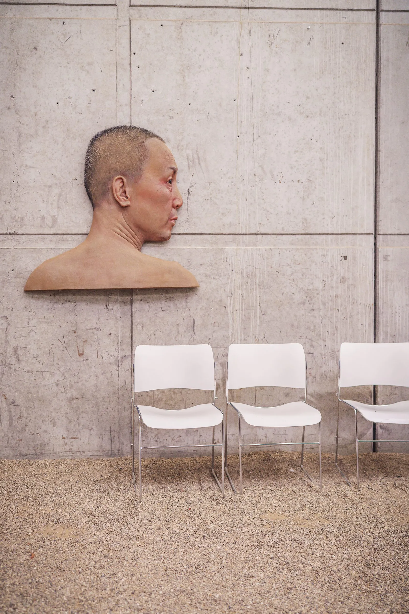 Portrait: a bust in profile on a concrete wall, three white chairs below
