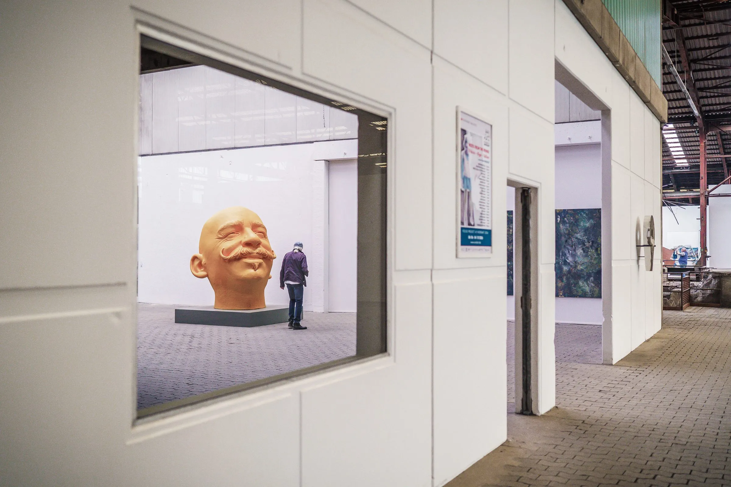 Through a window in a white wall you see a huge head with a moustache, a visitor beside it