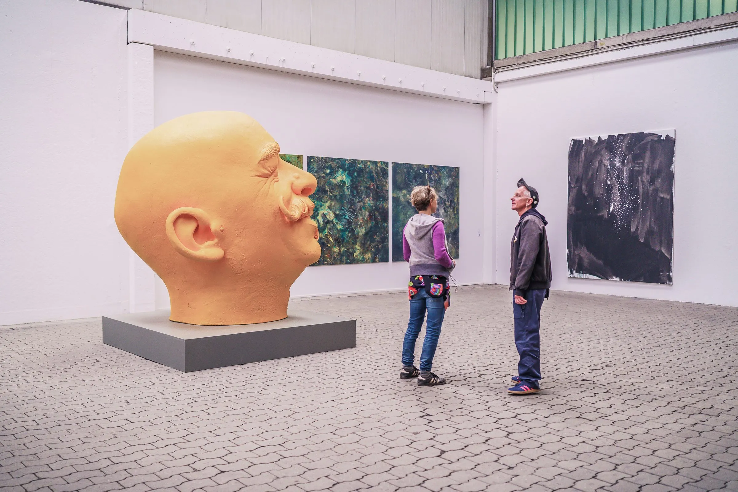 A huge head with a moustache on a plinth, two visitors stand in front and look up